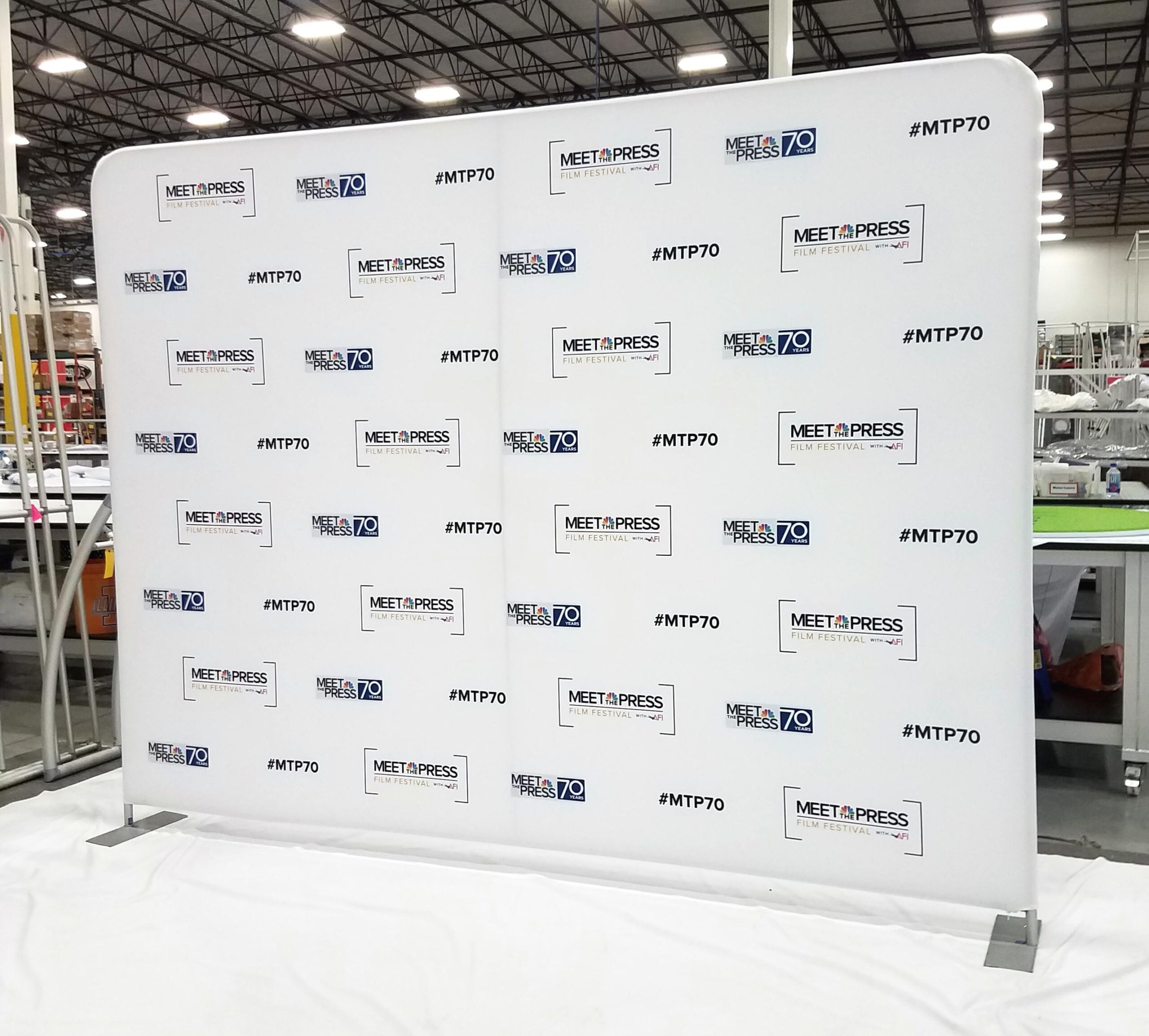 A step and repeat banner with logos for the Meet the Press film festival, NBC News 70th anniversary, and hashtag #MTP70, set up in a large indoor space with industrial ceiling and surrounding tables and shelves.