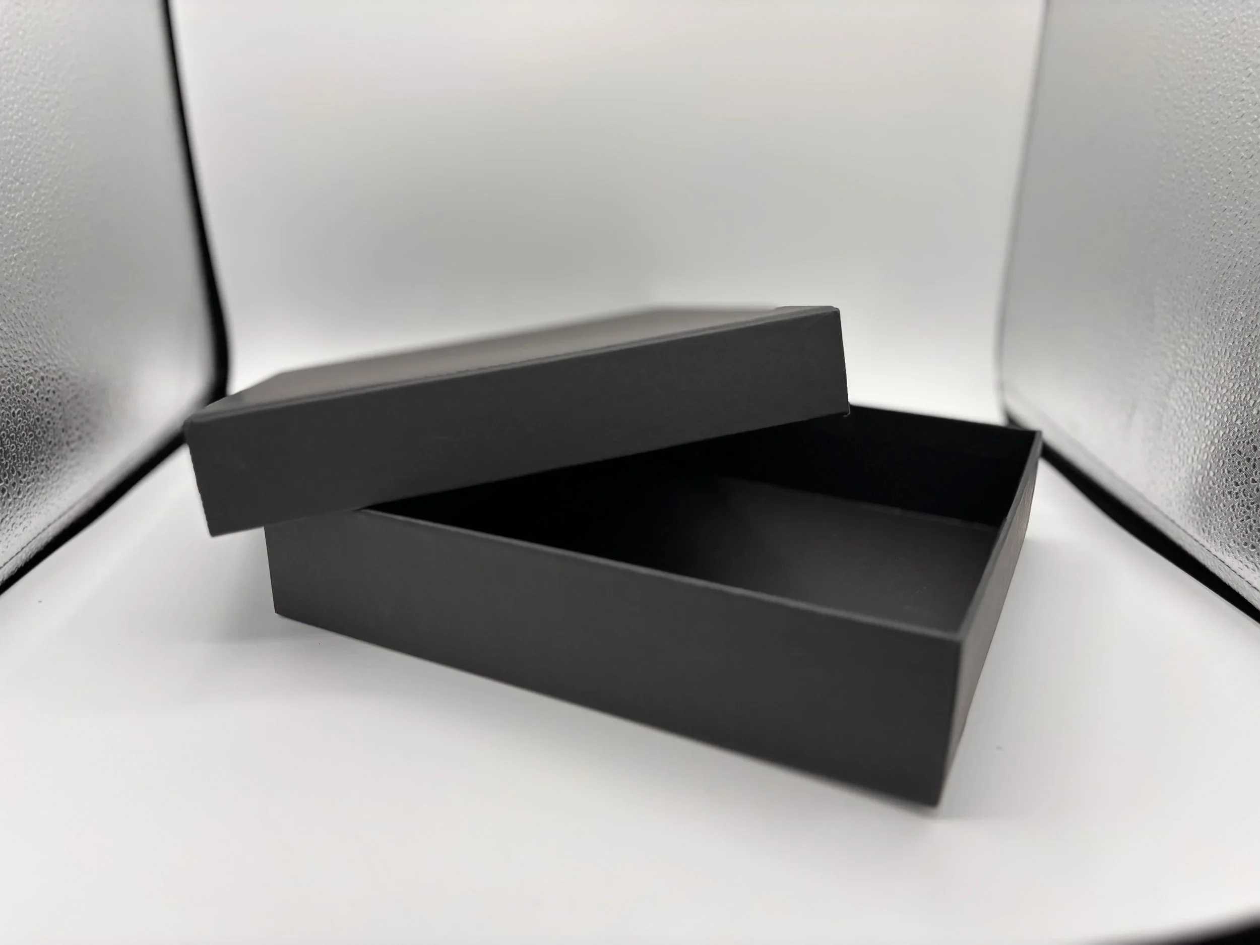 Open black gift box with a removable lid on a white surface, with a silver textured backdrop.