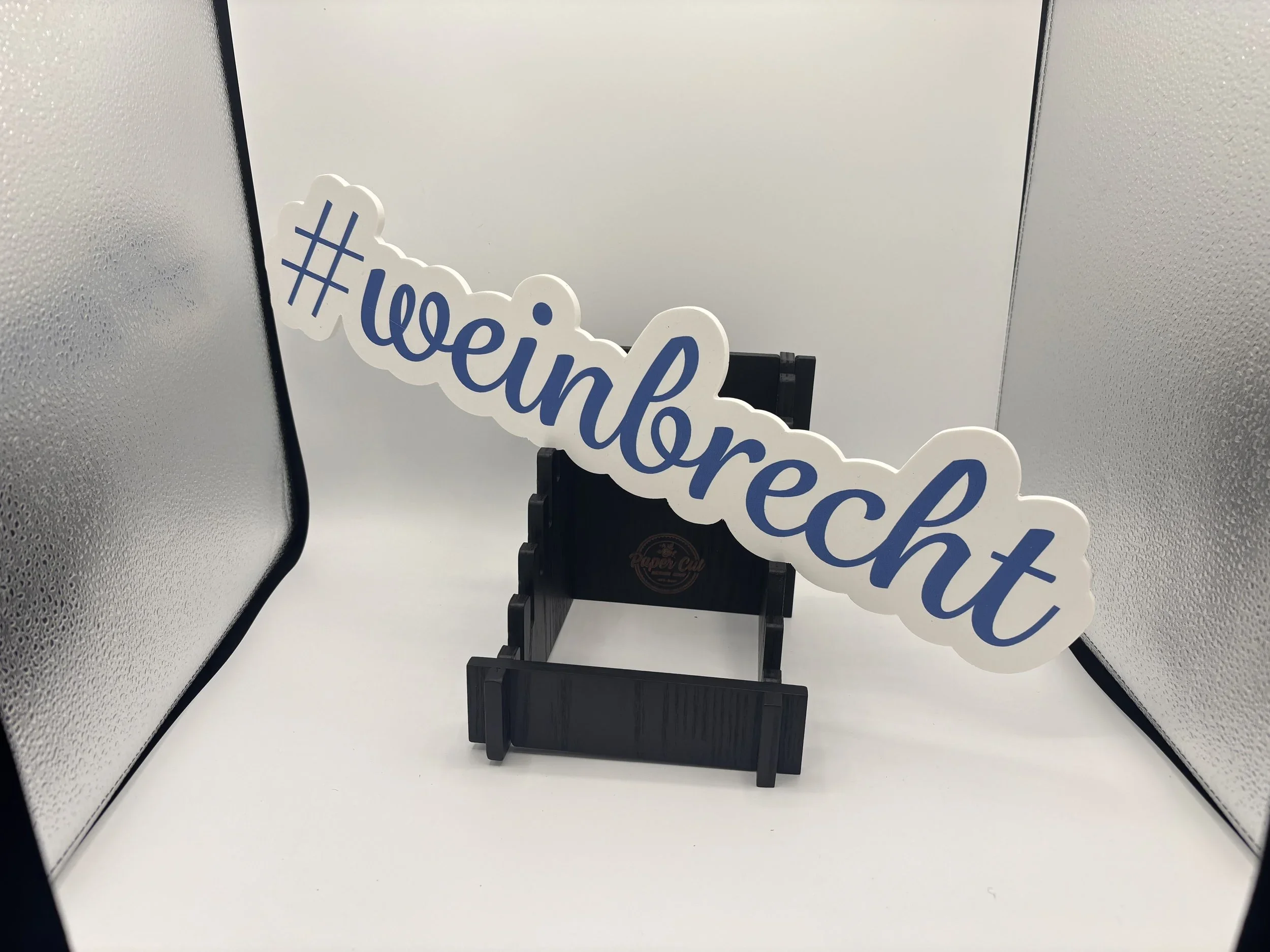 A sign with the hashtag #weintrecht placed on a black easel, photographed in a white and metallic background setup.