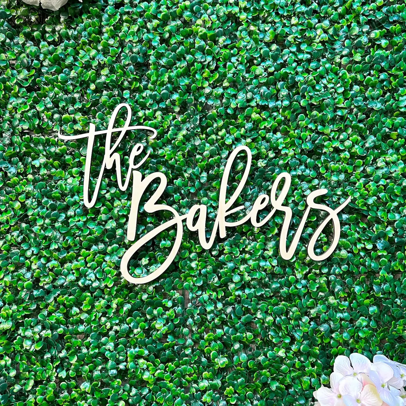 A lush green wall with a neon sign that says 'the Bakery' in cursive font, and a cluster of white flowers at the bottom right corner.