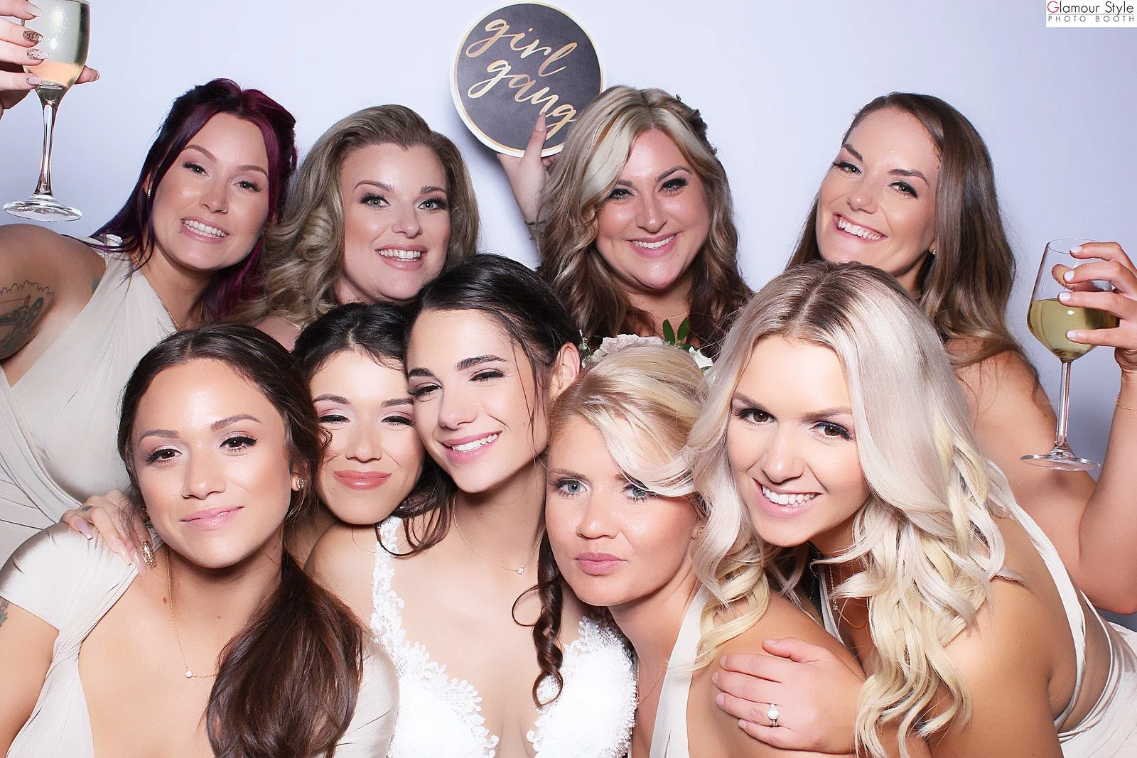 A group of women at a bridal shower, smiling and celebrating, with one holding a sign that says "girl gang" and two holding glasses of wine.