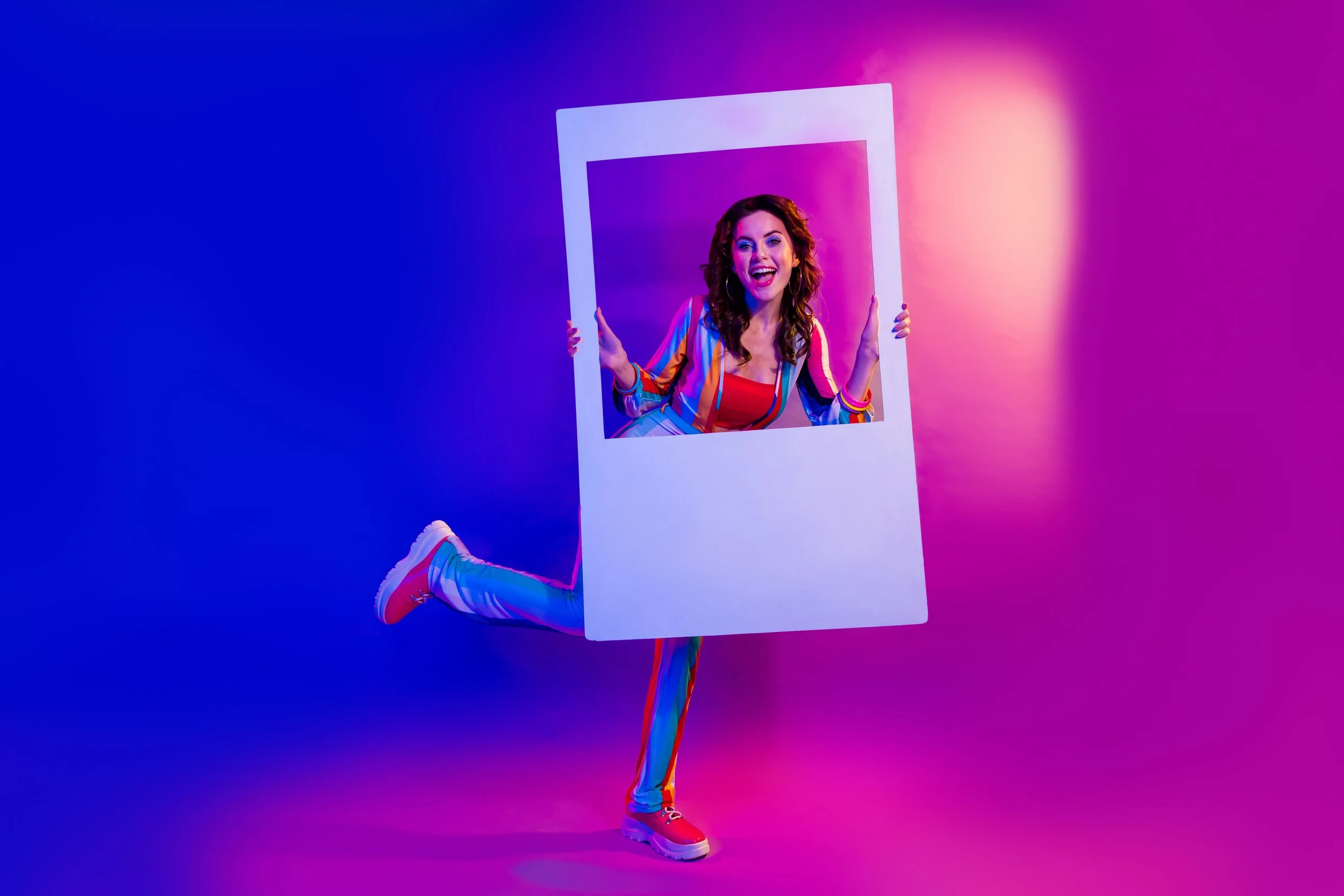 A woman in vibrant, rainbow-colored clothing holding a large white frame around her head, smiling, with one leg raised in front of a colorful gradient background.