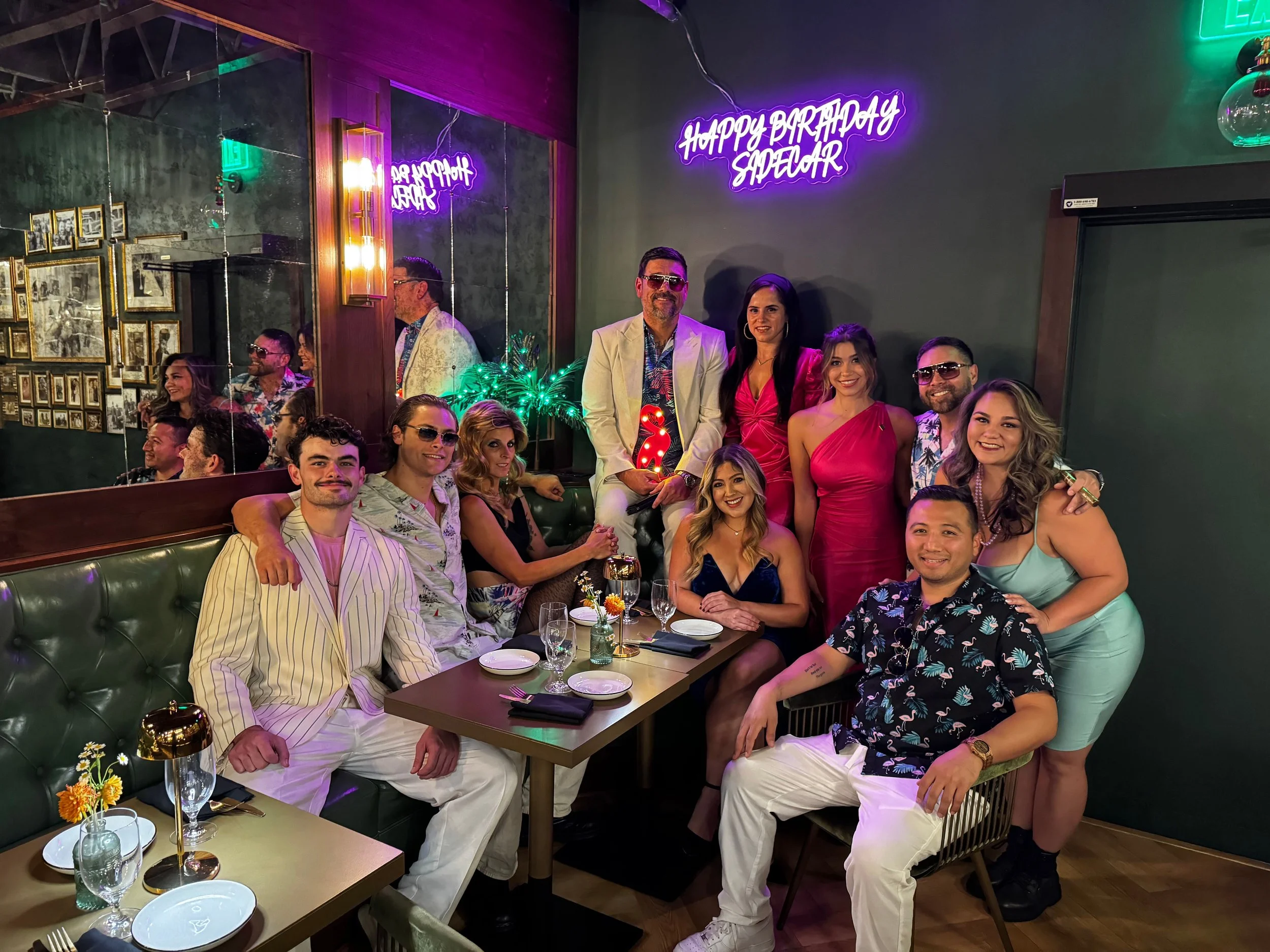 Group of people celebrating a birthday at a restaurant, with a neon sign that says 'Happy Birthday Sage Car' in the background. The group is dressed in colorful outfits, smiling and posing for the photo.