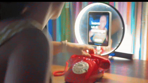 Person taking a selfie with a ring light, red vintage telephone, and a smartphone in front of a mirror.