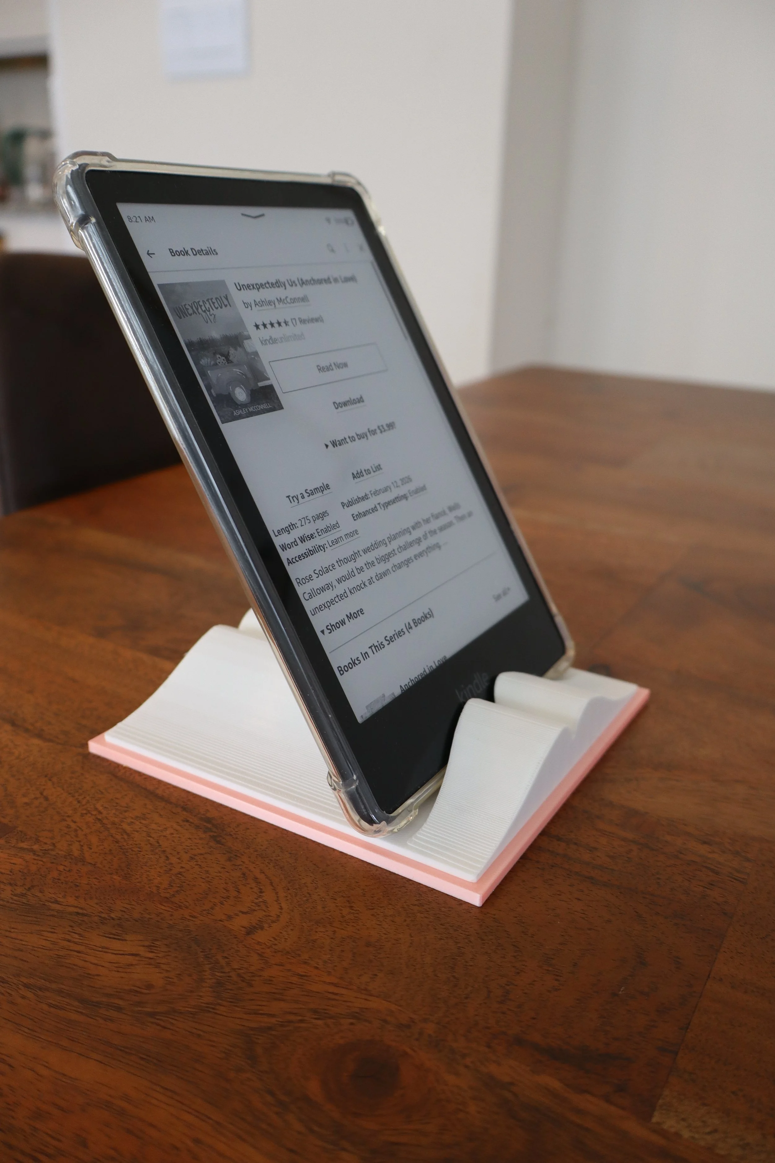A smartphone displaying the Kindle app with a book titled 'Unexpectedly Us' by Ashley McConnell on a pink and white stand on a wooden table.