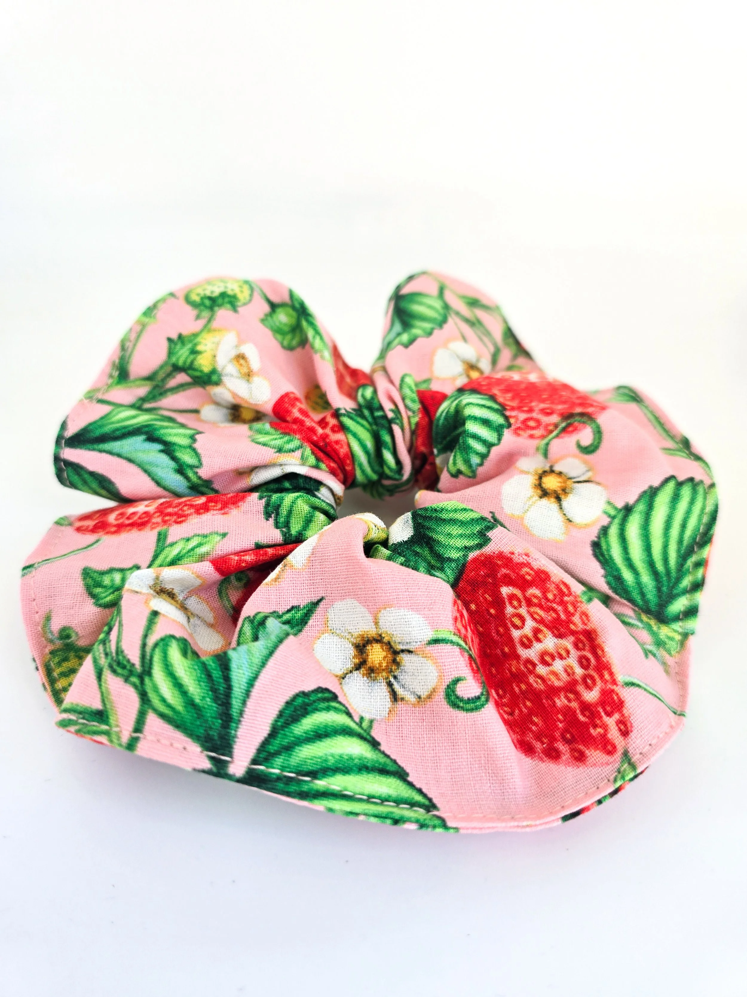 A pink fabric scrunchie with a strawberry and green leaf print on a white background.