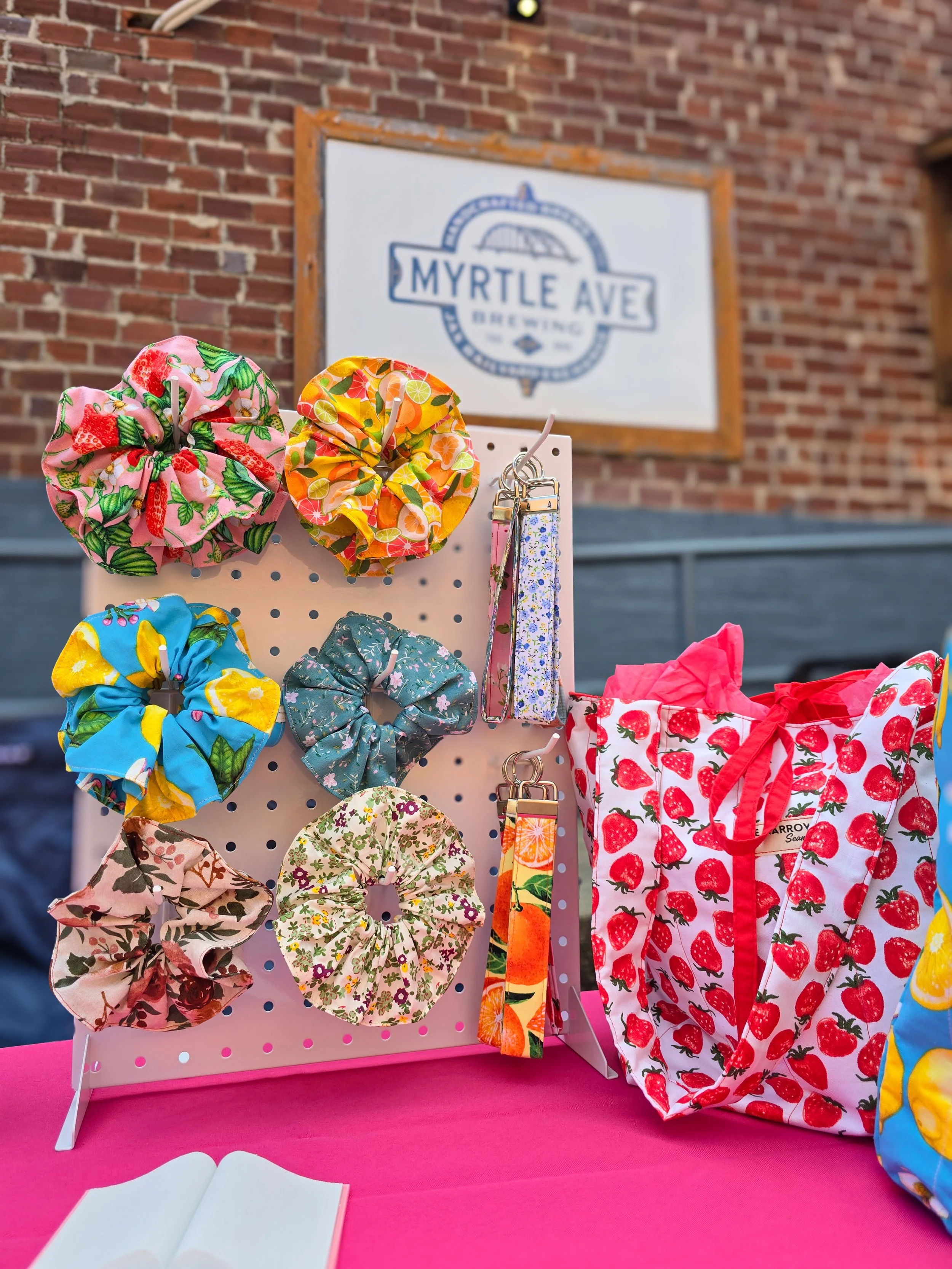 Colorful fabric scrunchies displayed on a white pegboard at an outdoor market, with a brick building and a sign reading 'Myrtle Ave Brewing' in the background, and a pink tablecloth in the foreground.