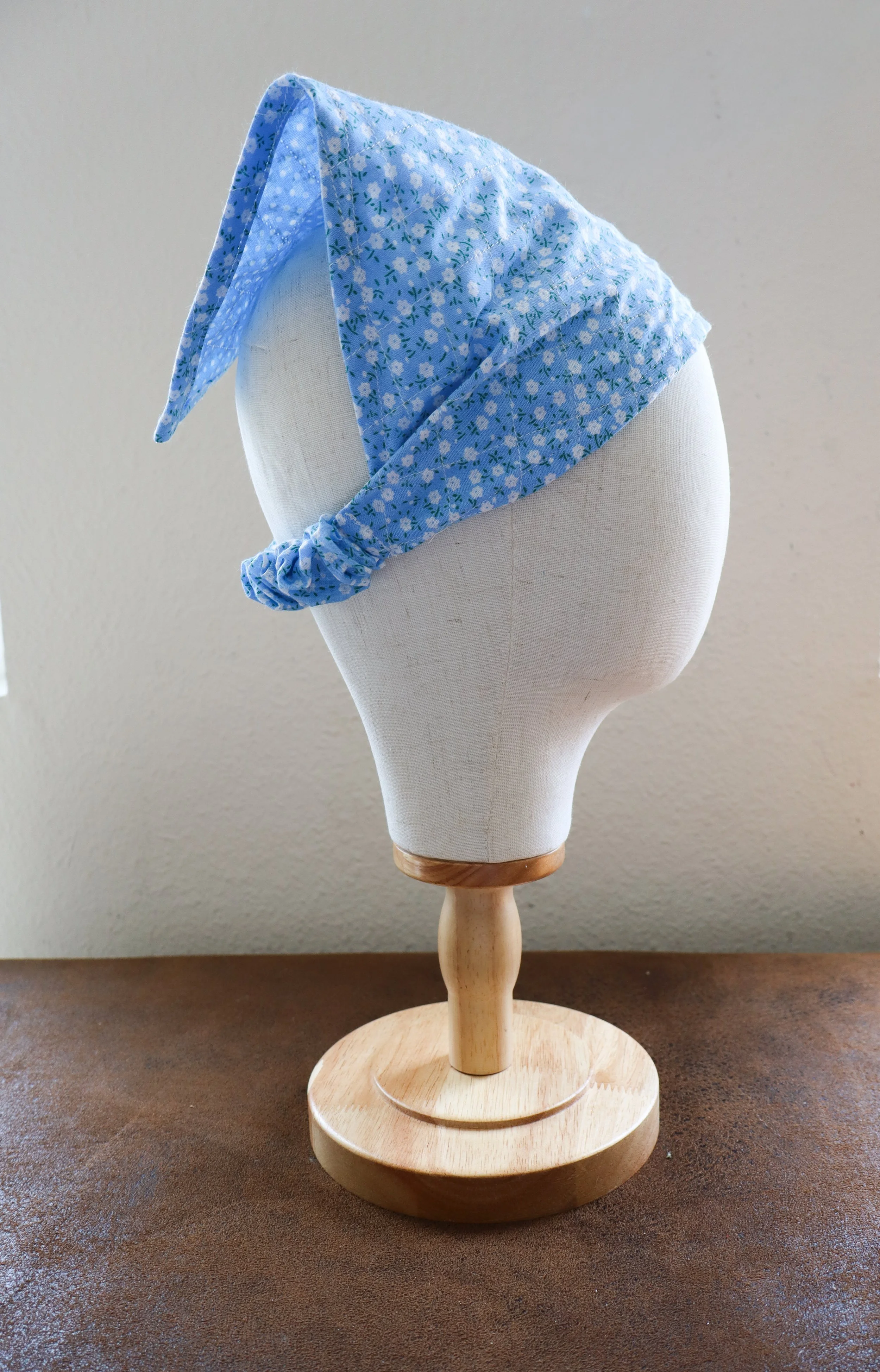 Blue floral-patterned fabric headscarf displayed on a mannequin head with a wooden base on a brown surface.