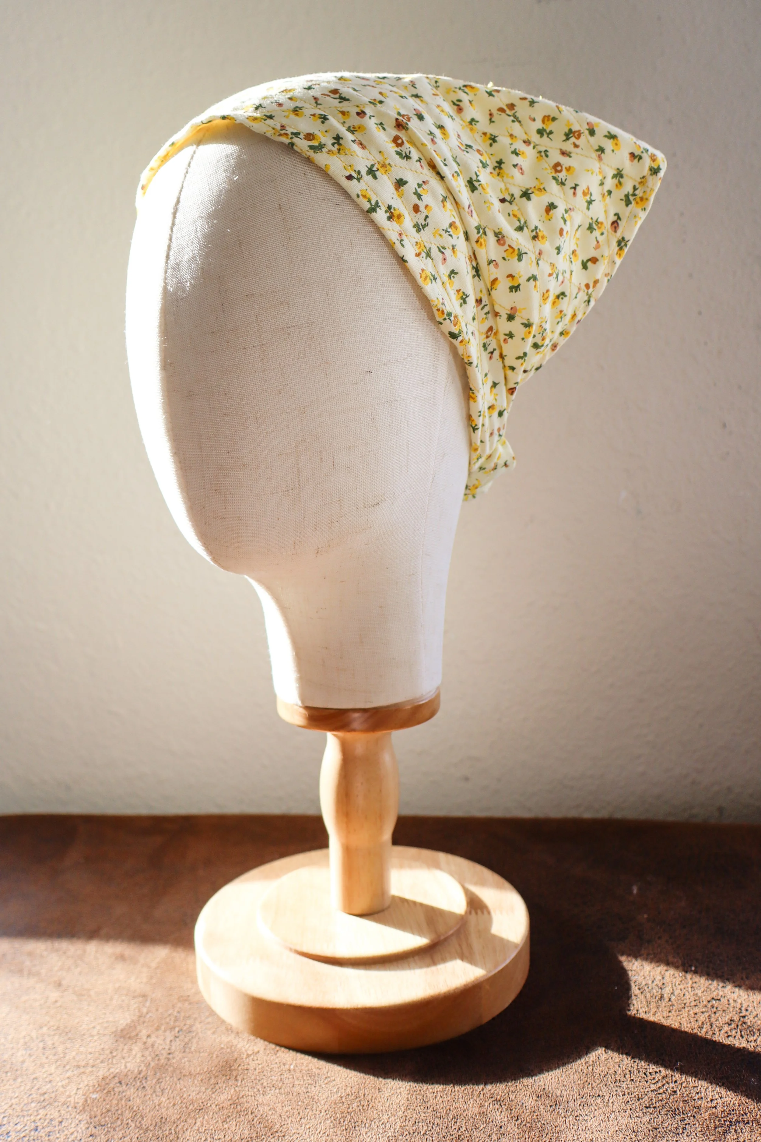 Wooden mannequin head with fabric bonnet with small yellow, orange, and green floral pattern, placed on a dark brown surface against a plain wall.