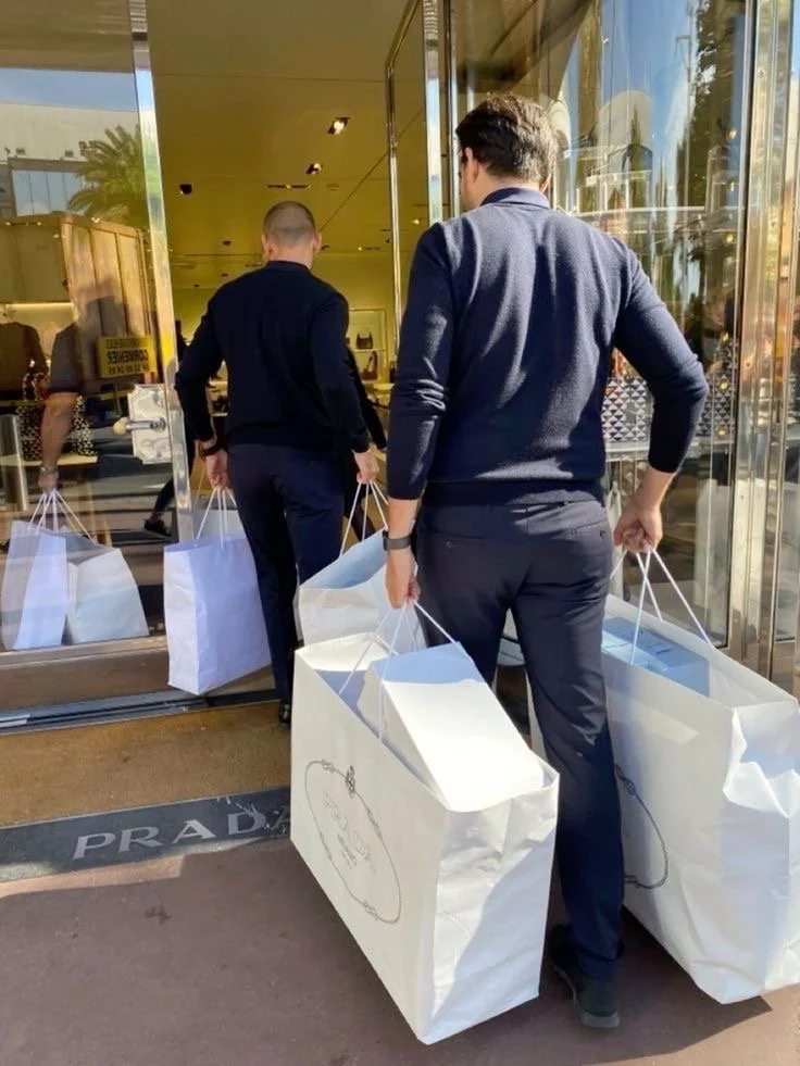 Two people in dark clothing carrying large shopping bags while entering a luxury store, with a reflection of the store's interior and palm trees visible in the glass door.