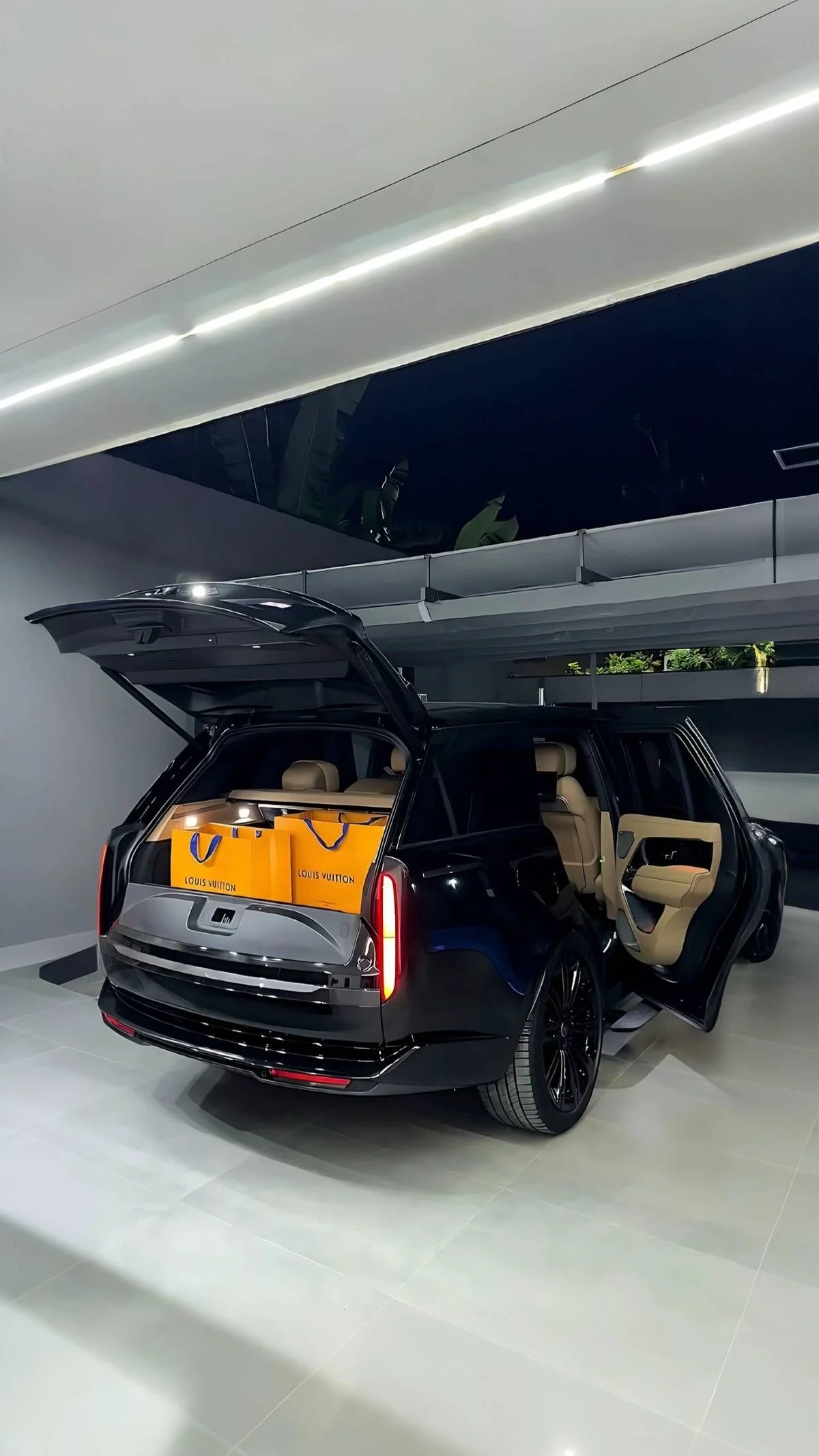 A black luxury SUV with an open rear hatch showing orange Louis Vuitton shopping bags inside. The interior is beige with open front passenger door, parked indoors with modern lighting and architecture.