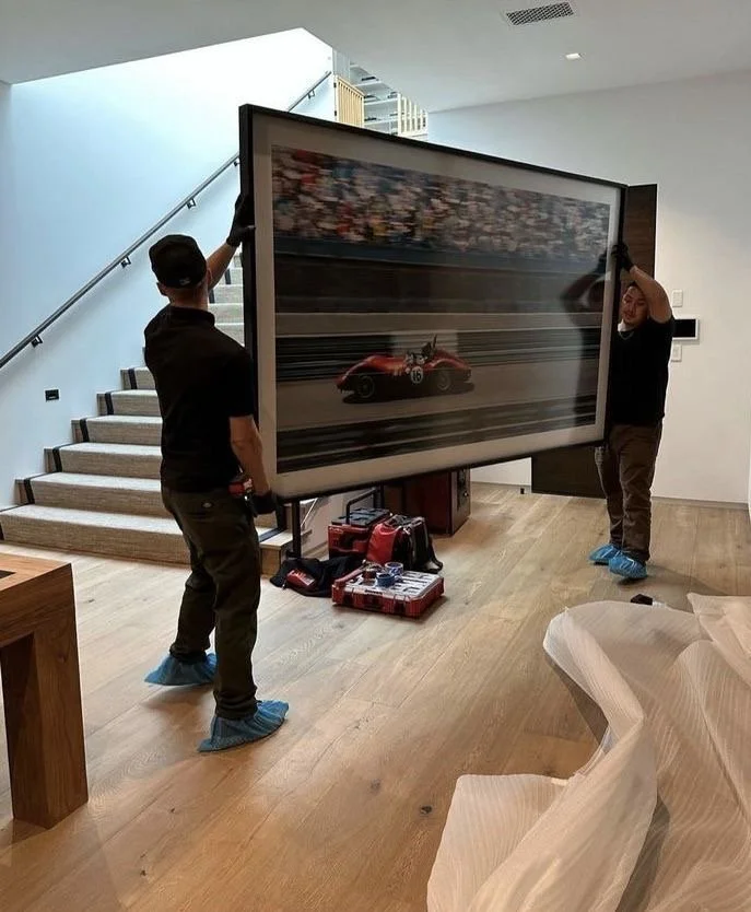 Two workers in shoe covers are installing a large framed art piece of a racing car on a wooden floor indoors. One person is holding the top side, and the other is adjusting the side of the frame. The art depicts a red race car on a track with a blurred crowd in the background.