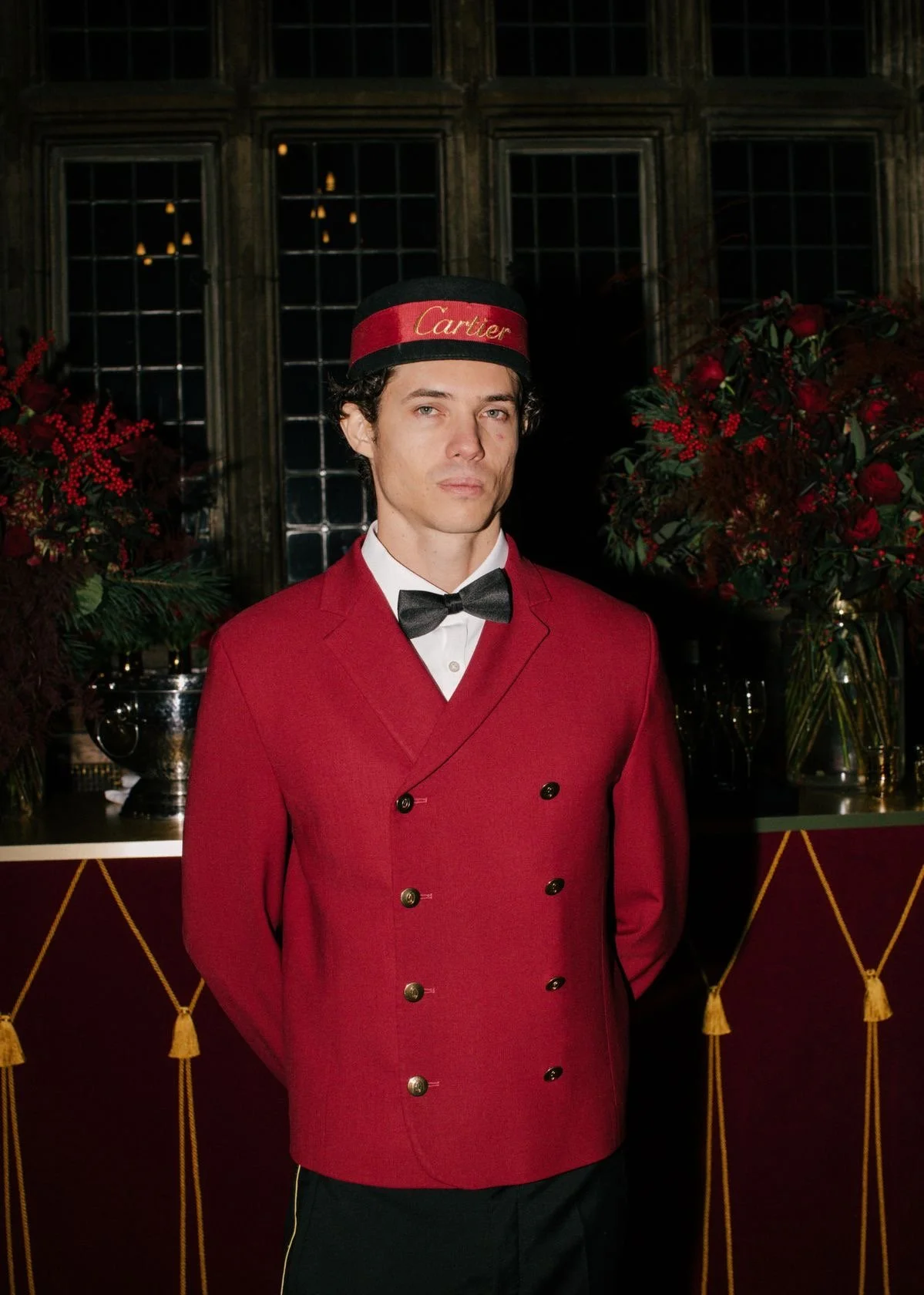 A man in a red double-breasted jacket, white shirt, black bow tie, and a capsule hat with the word 'Cartier' on it, standing indoors with floral arrangements in the background.