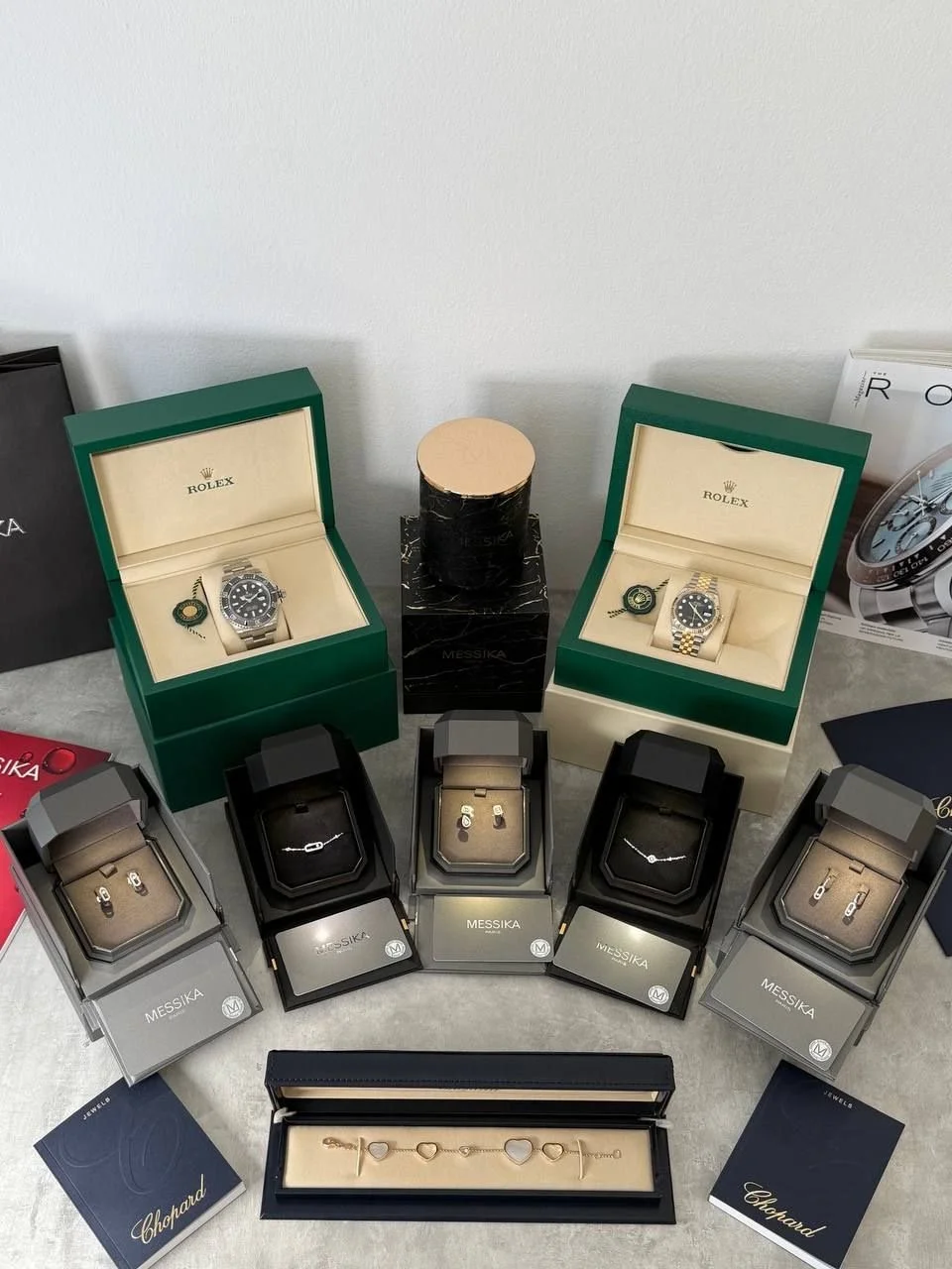 Display of luxury watches, jewelry, and accessories, including two Rolex watches in green boxes, several other watch boxes, and a jewelry bracelet.