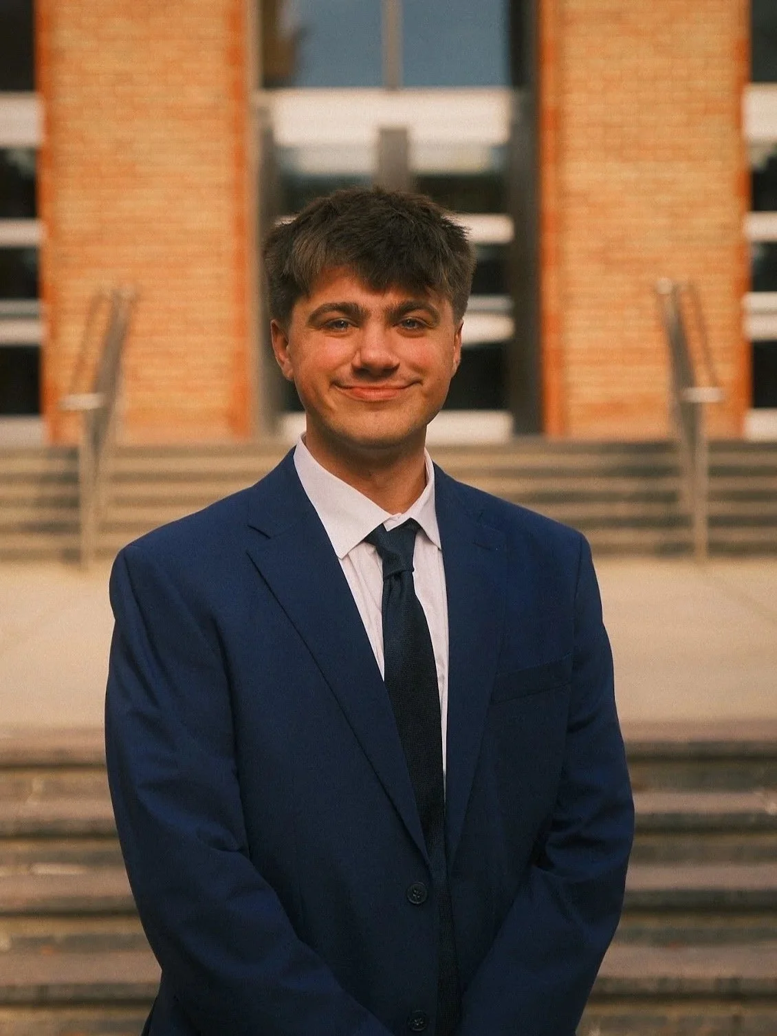 A young man with dark hair wearing a navy blue suit, white shirt, and dark tie, standing on steps in front of a building with brick columns and large windows, smiling at the camera.