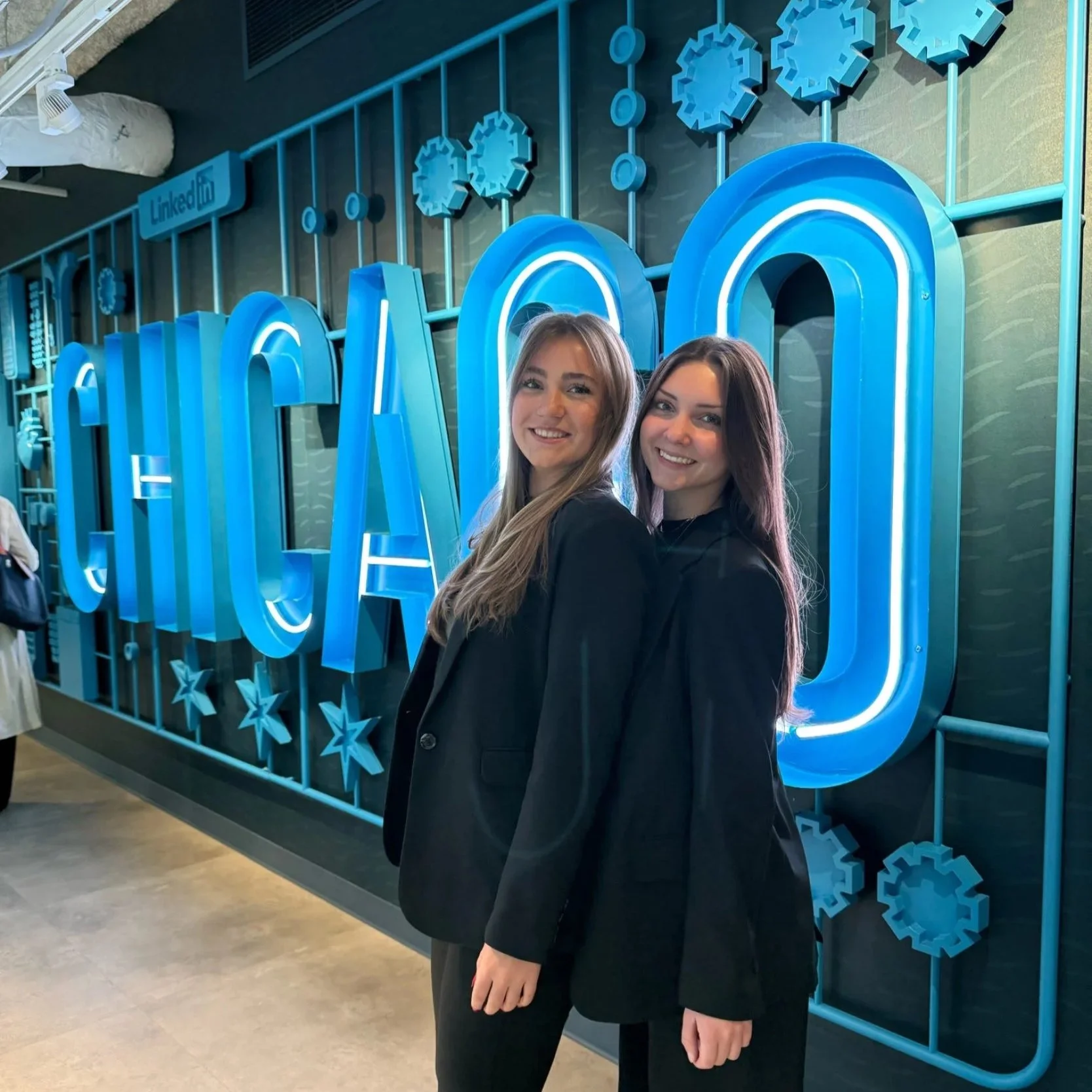 Two young women with blonde and brown hair smiling and standing together in front of a large illuminated LinkedIn sign with blue snowflake decorations.