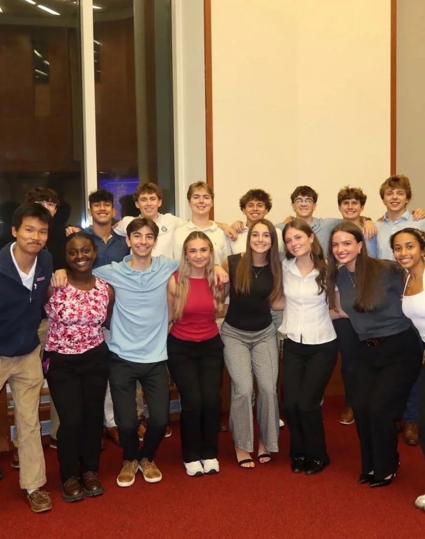 A group of young students, both boys and girls, posing together indoors at an event or gathering, smiling with arms around each other.