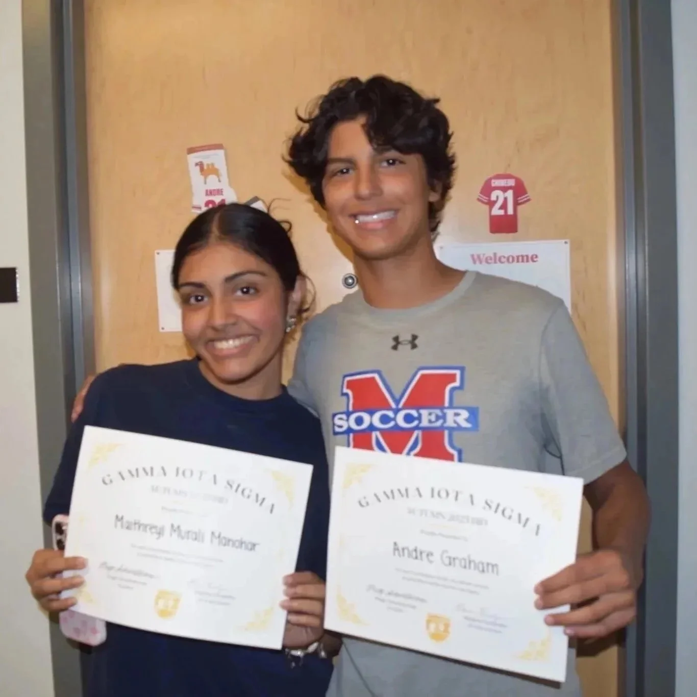 Two students holding certificates, standing inside a school hallway, smiling at the camera.