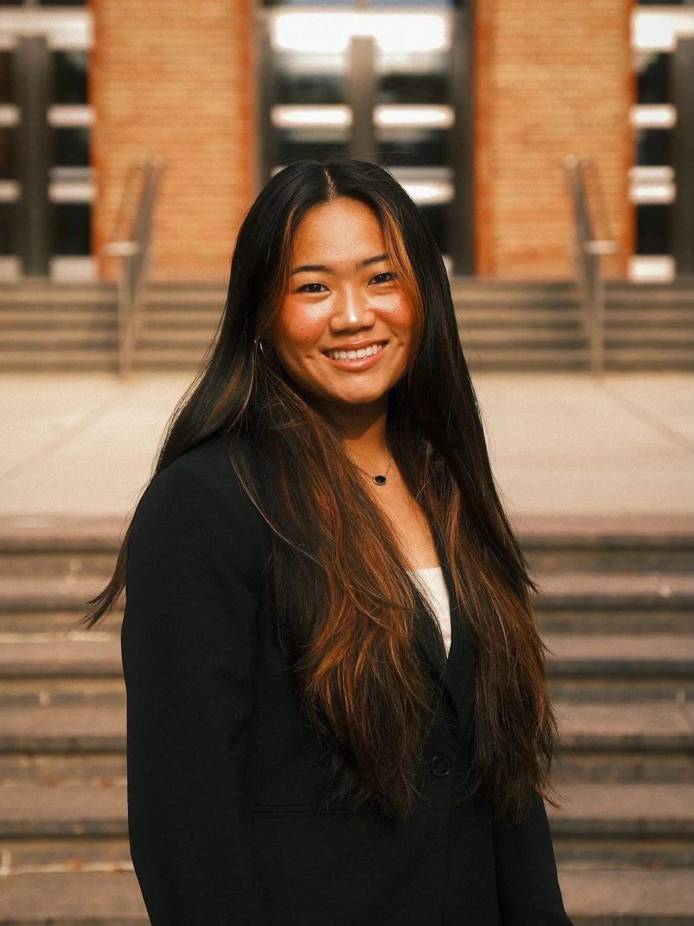 A young woman with long dark hair smiling, wearing a black blazer, standing in front of steps leading to a building.