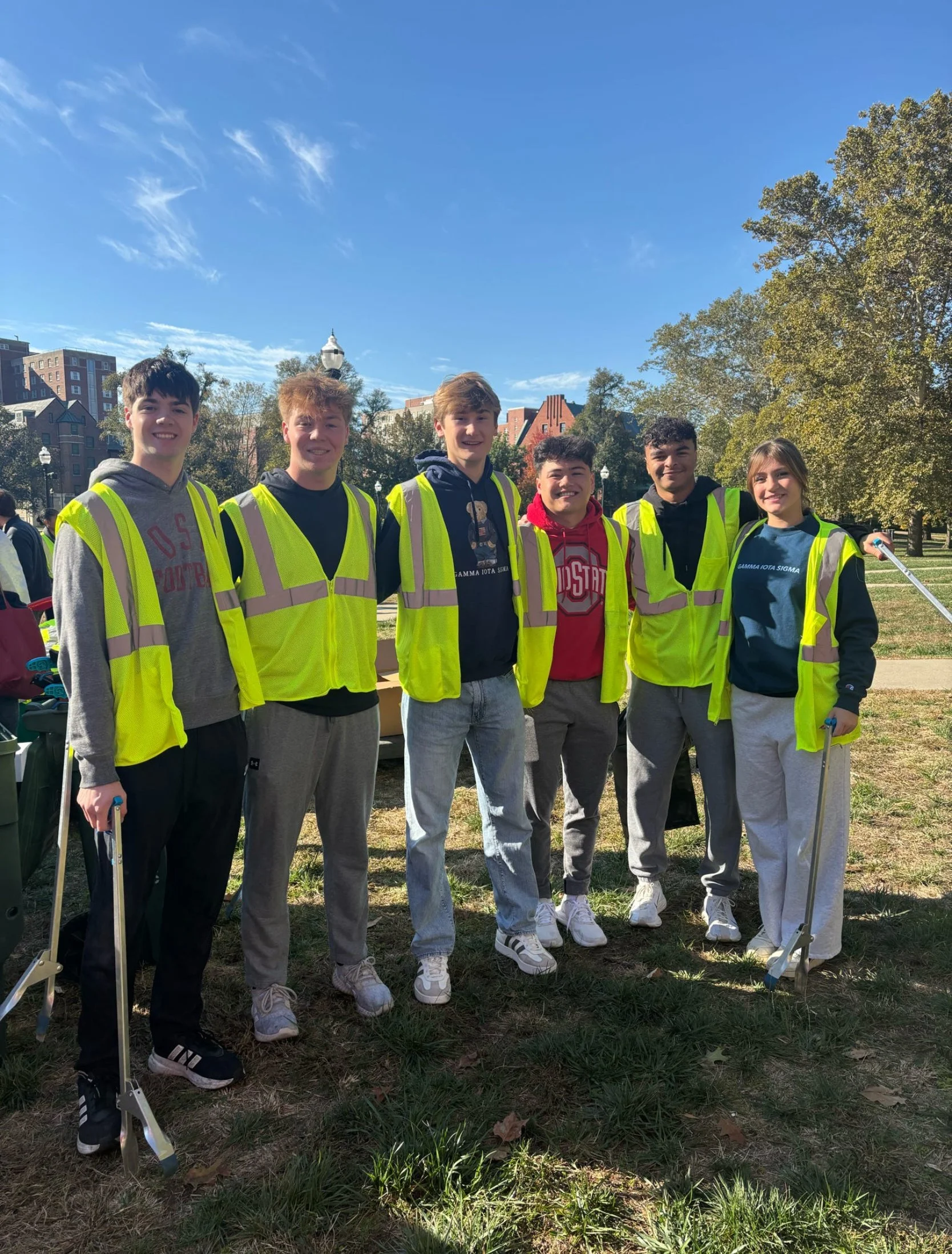 A group of six young people standing outdoors on a sunny day, wearing yellow safety vests. They are smiling and posing for the photo, with a park, trees, and a blue sky in the background.