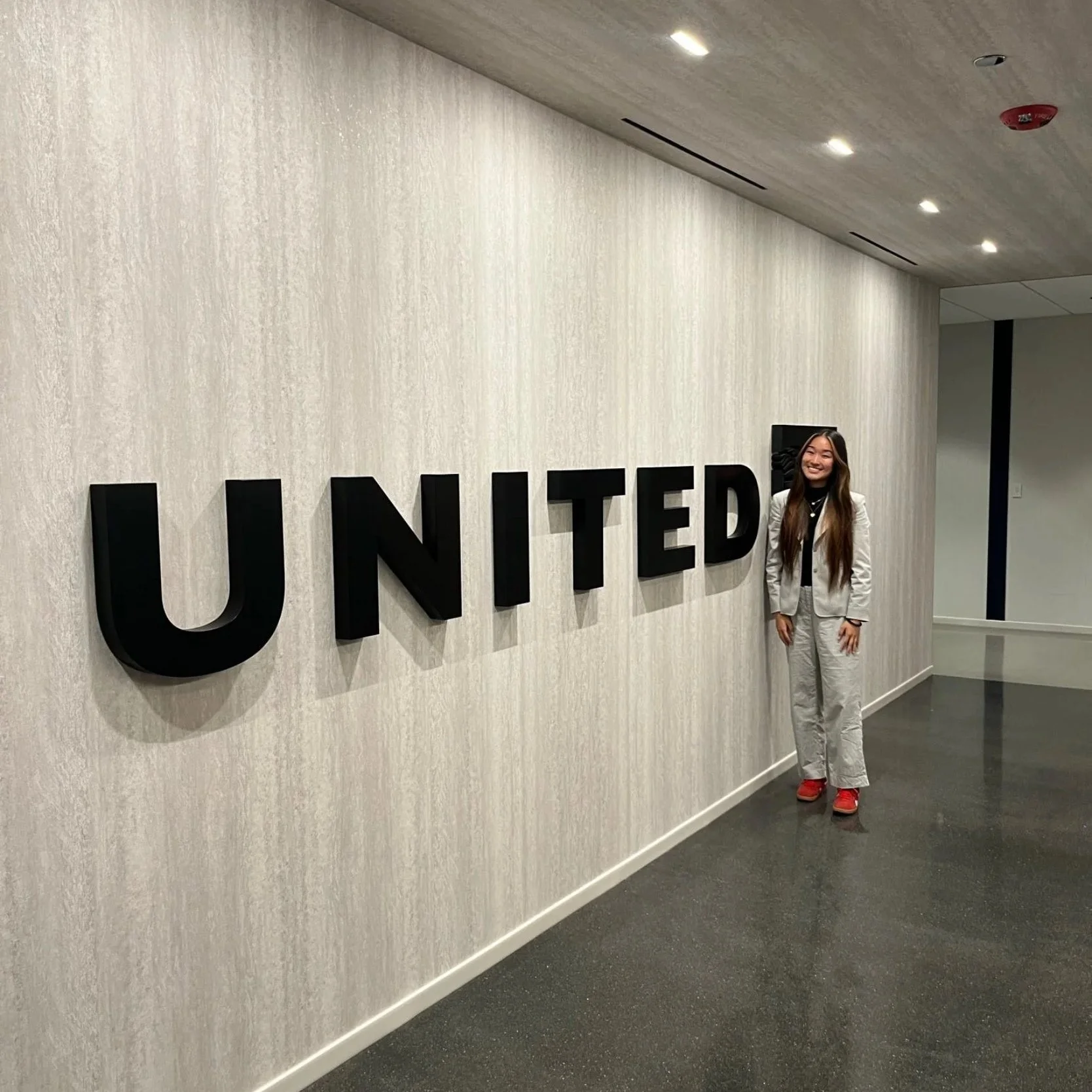 A woman with long brown hair wearing a gray suit and red shoes standing next to large black letters spelling 'UNITED' on a white wall in an indoor setting.