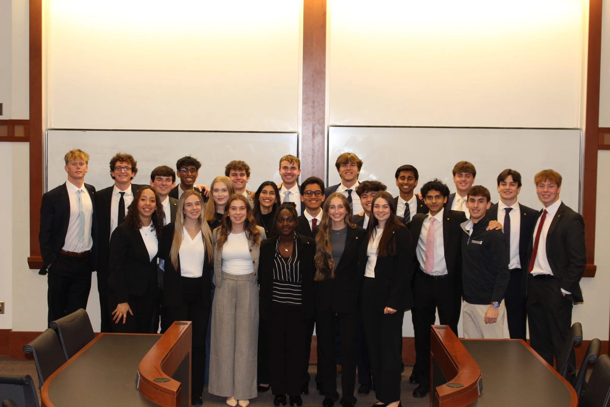 Group of young adults, dressed in formal business attire, posing together in a conference room with whiteboards behind them.
