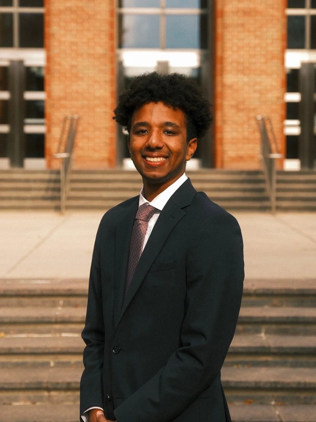 A young man in a suit standing outdoors in front of a brick building with stairs, smiling at the camera.