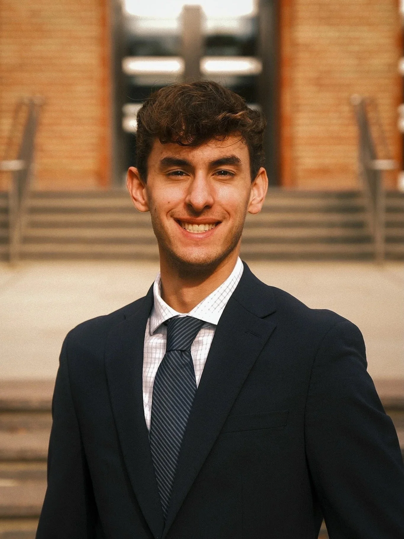 Young man in a business suit smiling in front of a brick building with stairs.
