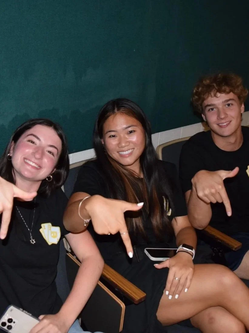 Three young adults sitting in chairs, smiling, and pointing at each other, wearing black shirts, in a room with dark green walls.