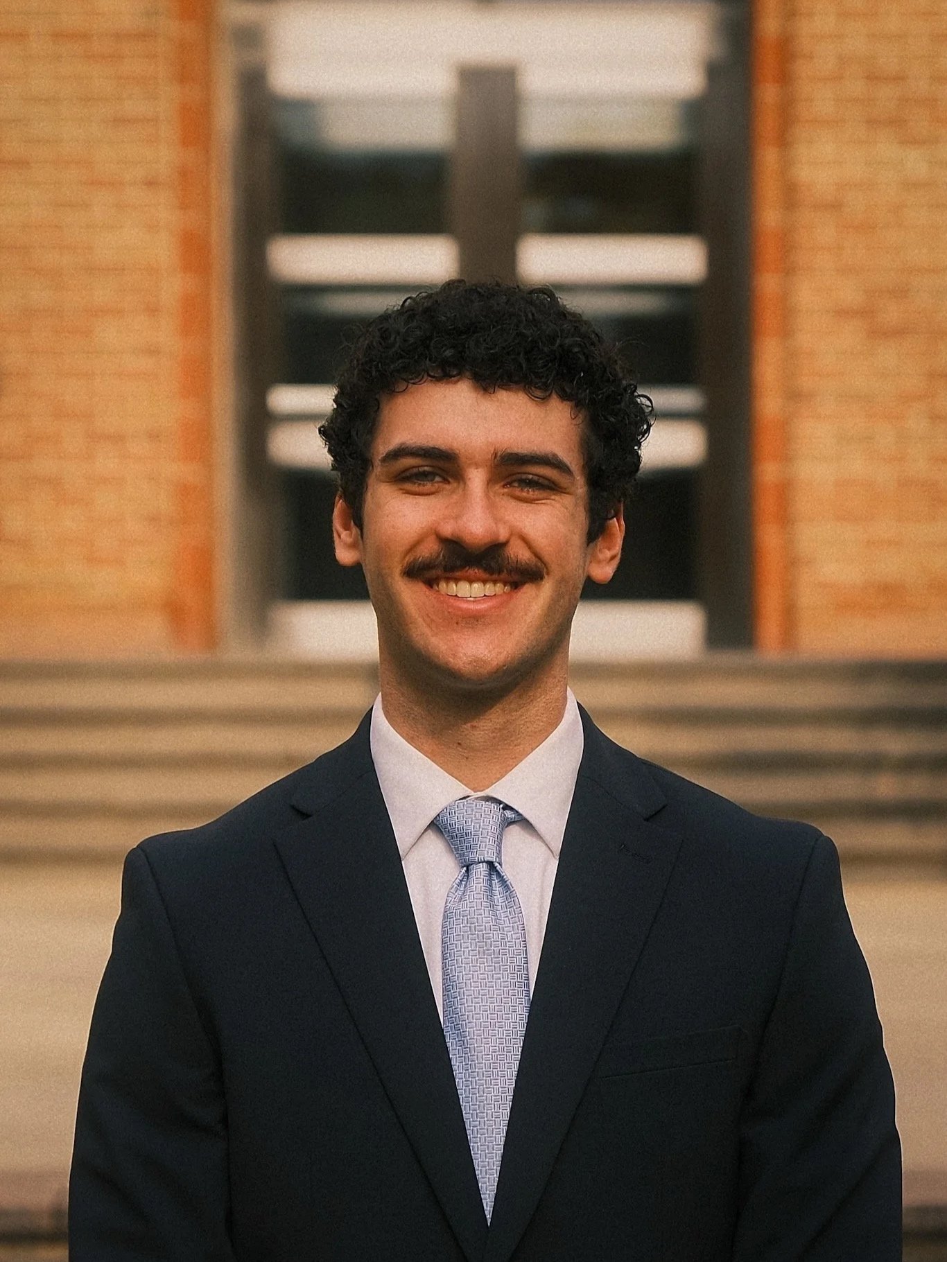 Young man in a dark suit and light blue tie standing outside in front of stairs and a building.