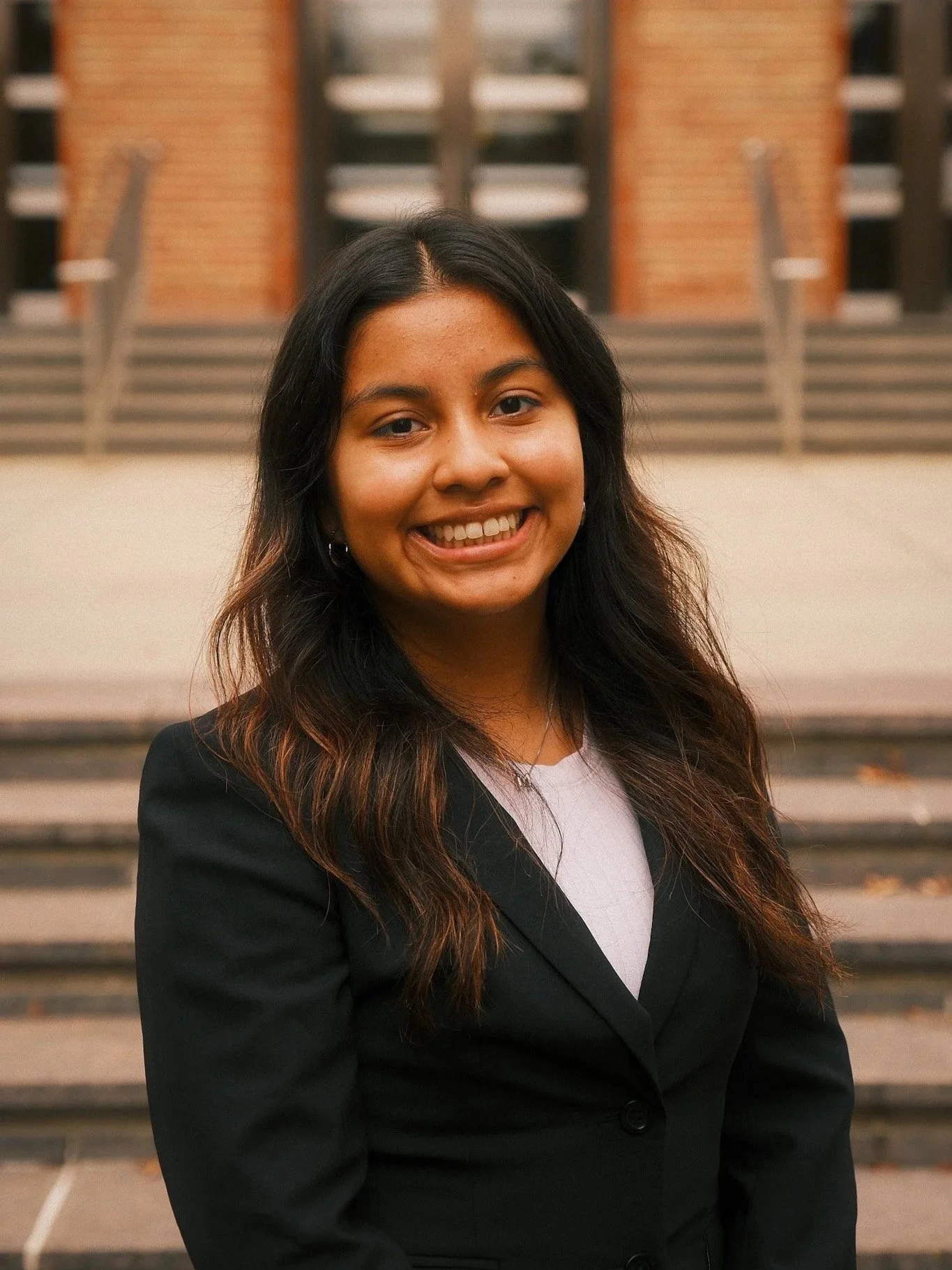 A smiling young woman with long dark hair wearing a black blazer standing outdoors in front of stairs and a building.