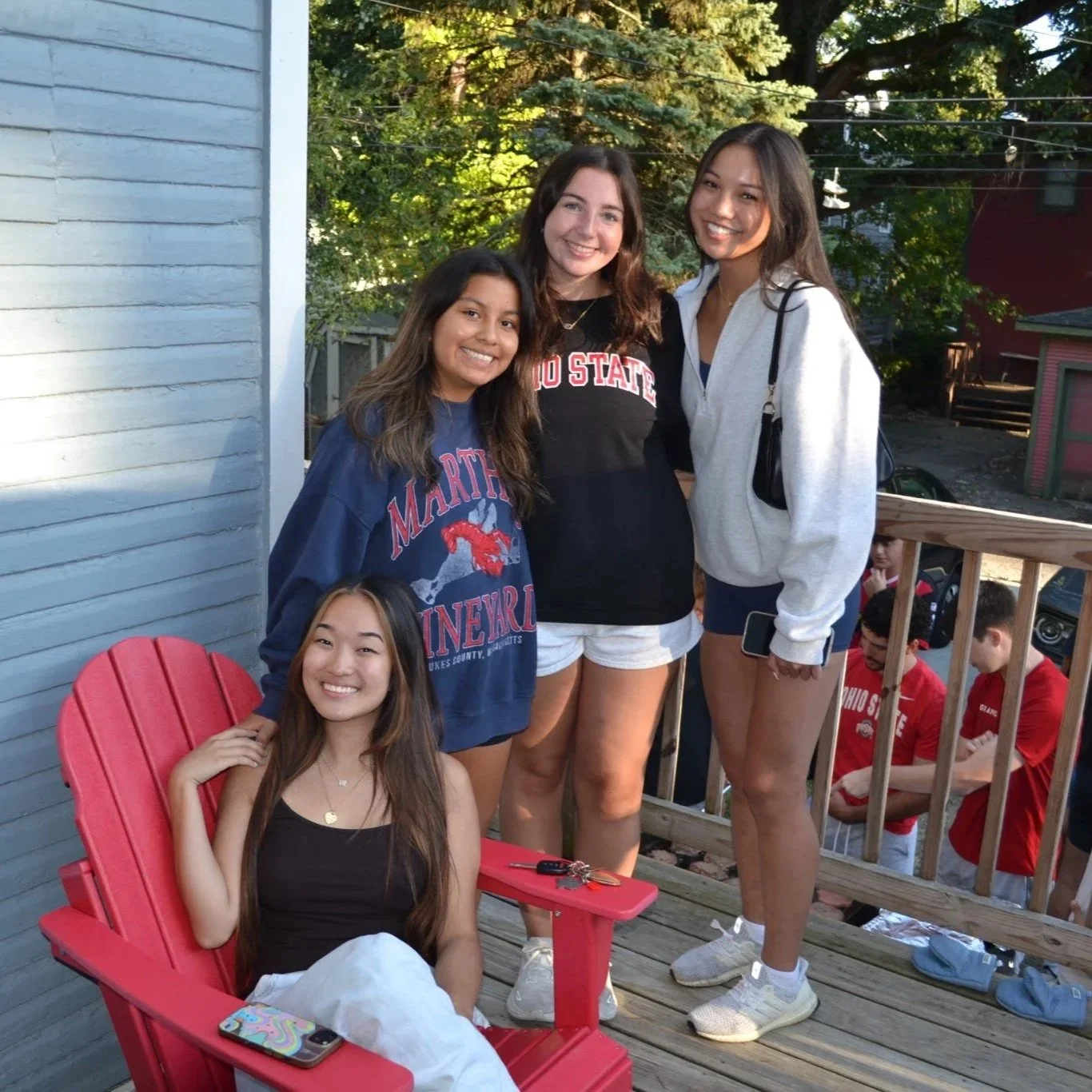 Group of five young women posing together outdoors on a wooden deck, with trees and houses in the background. Some are standing, one is sitting in a red chair, and they are smiling.