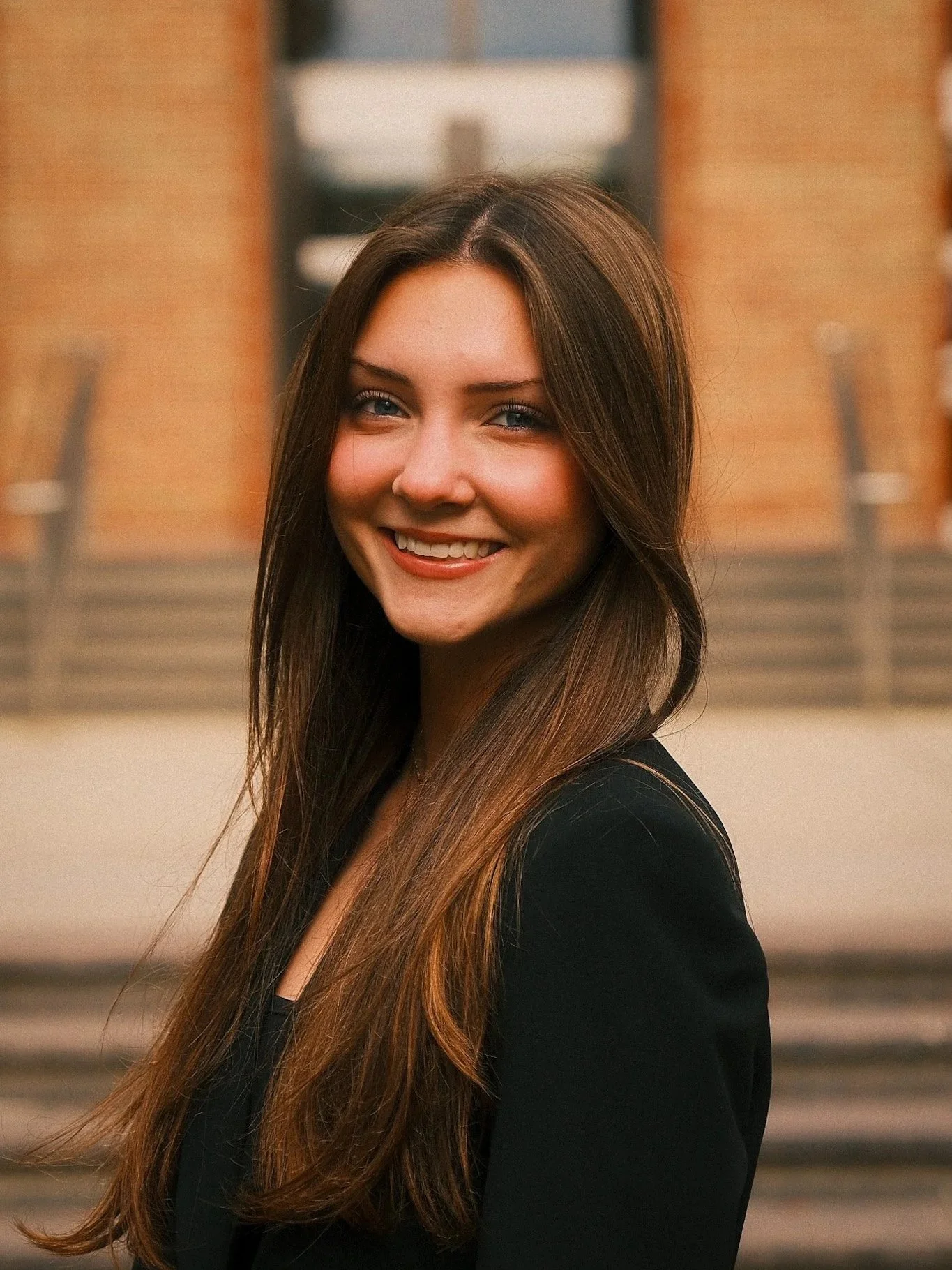 Young woman with long brown hair smiling outdoors with a brick and glass building in the background.