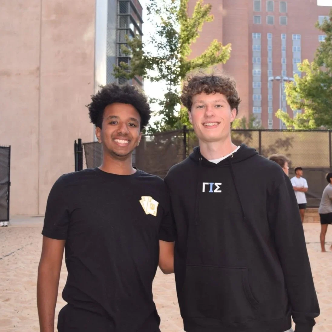 Two young men smiling, standing outdoors in front of a building with trees, one wearing a black T-shirt with a Greek letter and the other wearing a black hoodie with Greek letters, at a recreational area with a sandy surface and a fence.