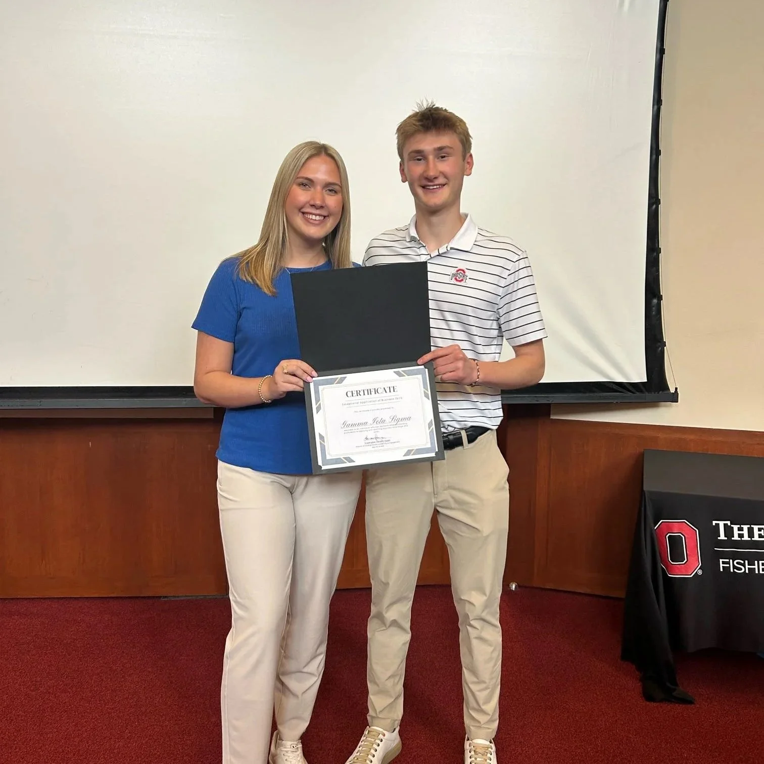 Young woman and young man standing together, smiling, holding a certificate and an open folder in front of a white wall and projector screen. The woman is dressed in a blue top and white pants, the man in a white striped polo and beige pants. There is a table with a black tablecloth that has a college logo and text on the right side.