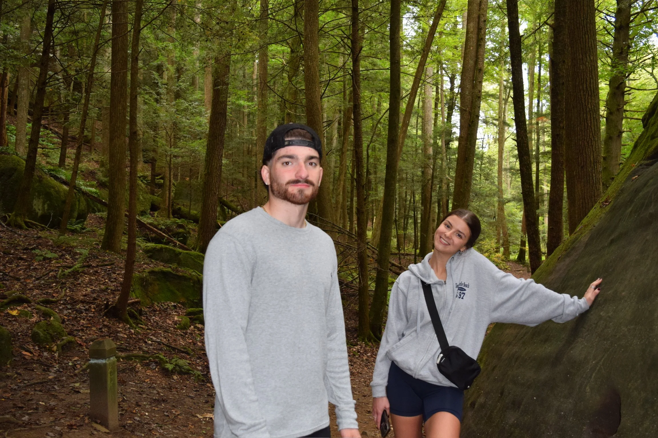 A man with a beard wearing a gray sweatshirt and a backward black cap and a woman in a gray hoodie and shorts smiling in a forest with tall trees and green foliage.