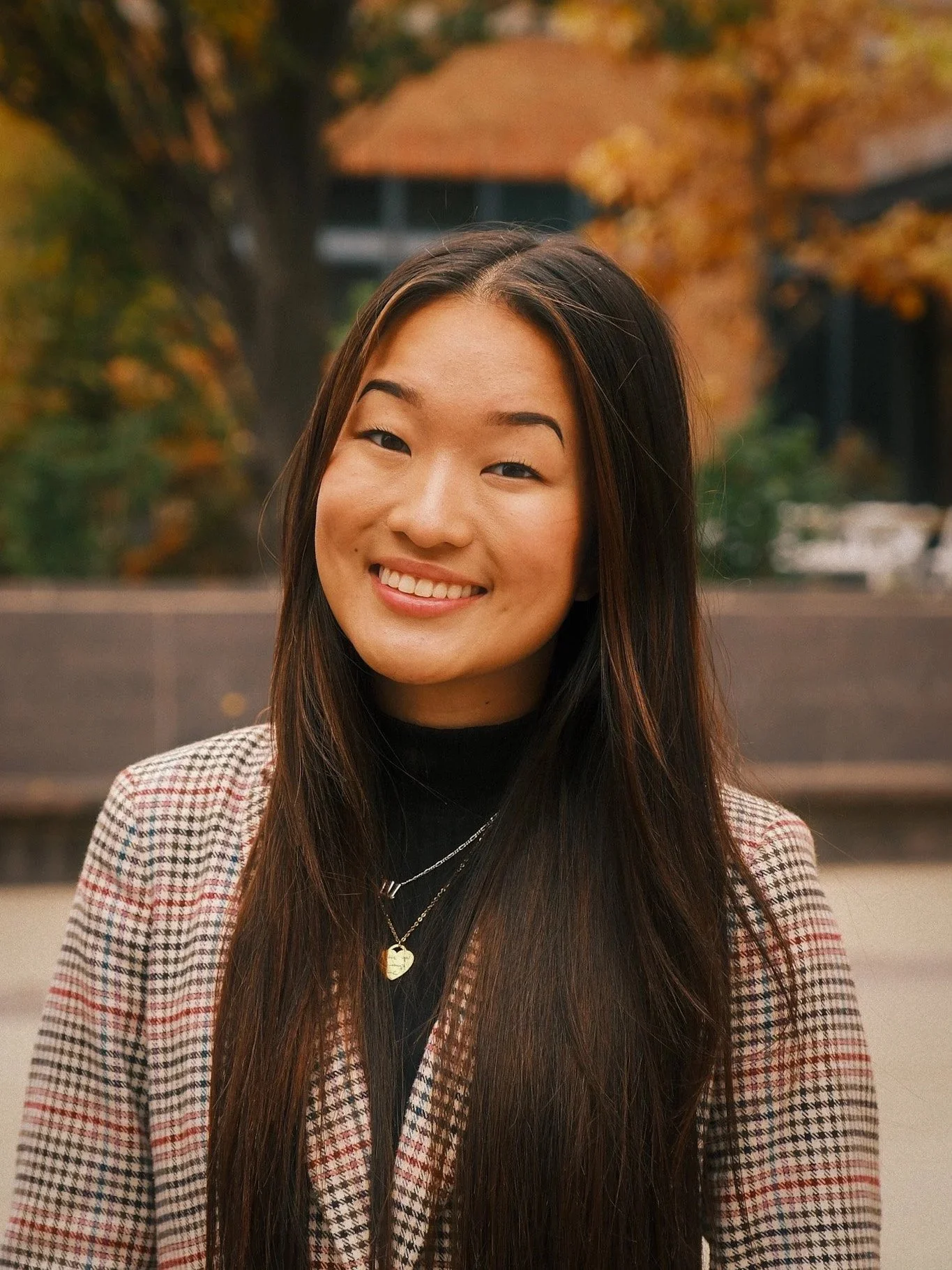 A smiling young woman with long dark hair wearing a black top, layered necklaces including one with a heart pendant, and a plaid blazer, standing outdoors with fall foliage in the background.