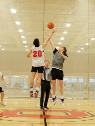 Two basketball players jumping for the ball at the start of a game, with a referee standing between them on an indoor court.