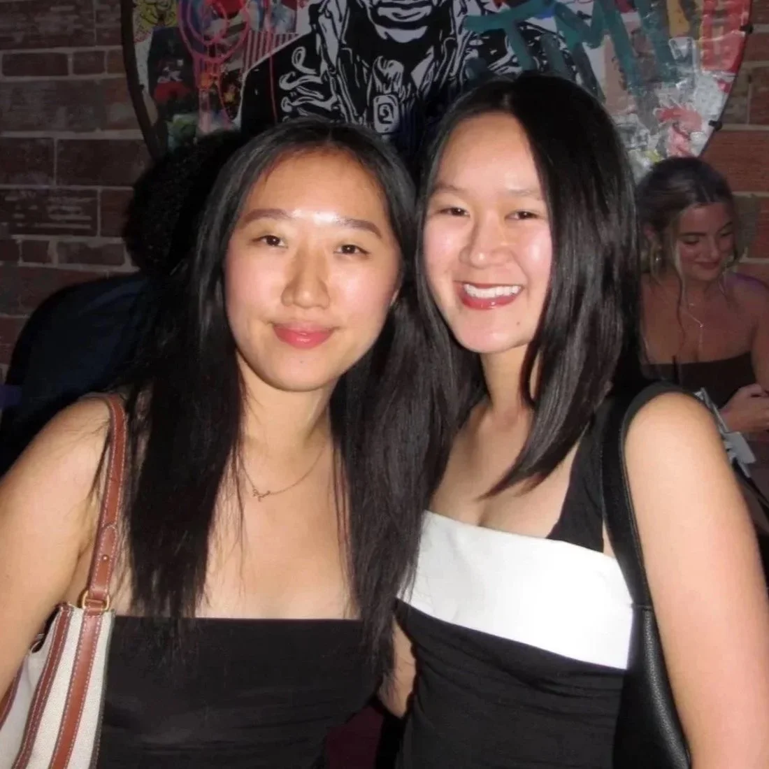Two women smiling, posing for a photo indoors. Behind them, a woman is looking at her phone, and there is brick wall decor.
