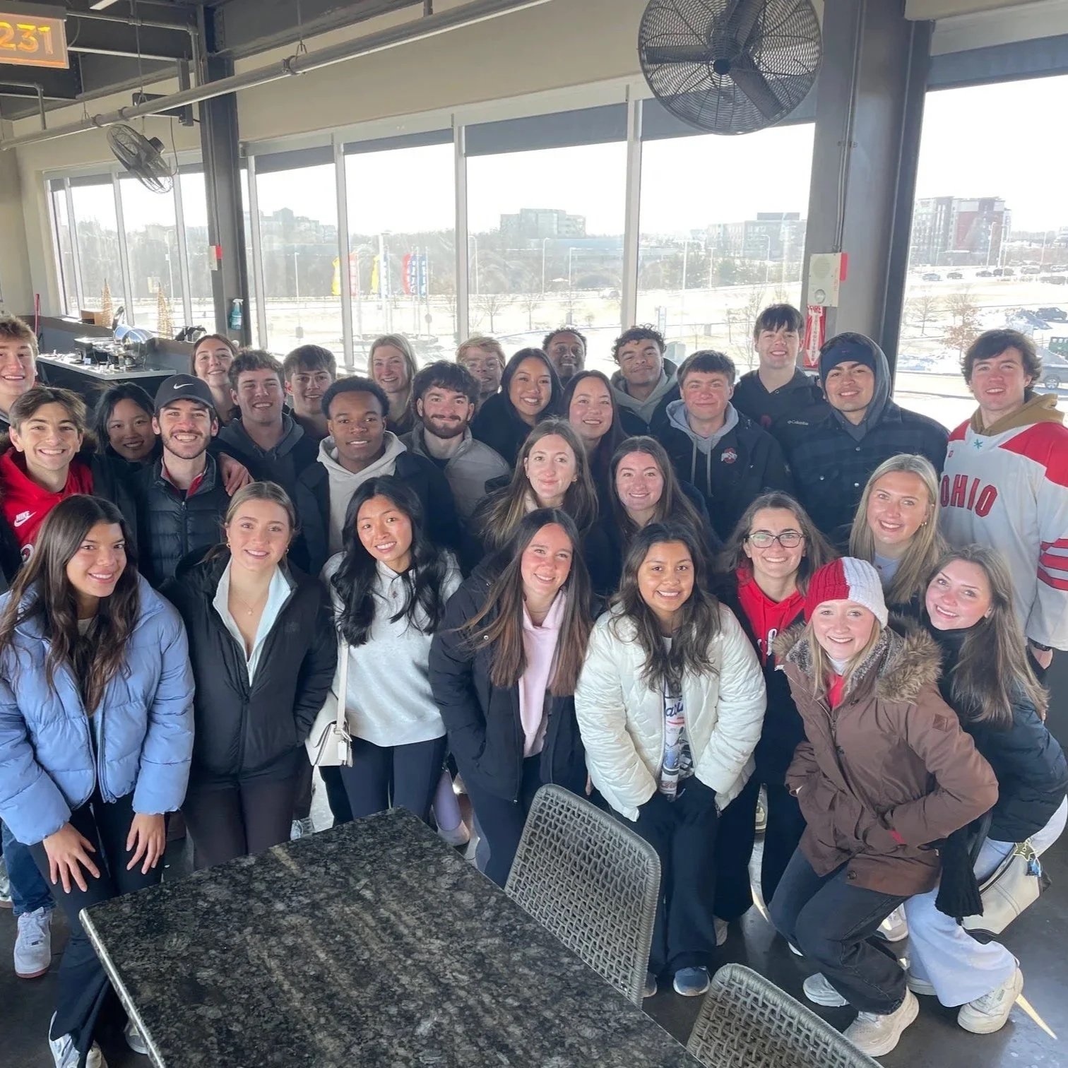 A group of smiling young people posing indoors near large windows with a snowy landscape and buildings outside.
