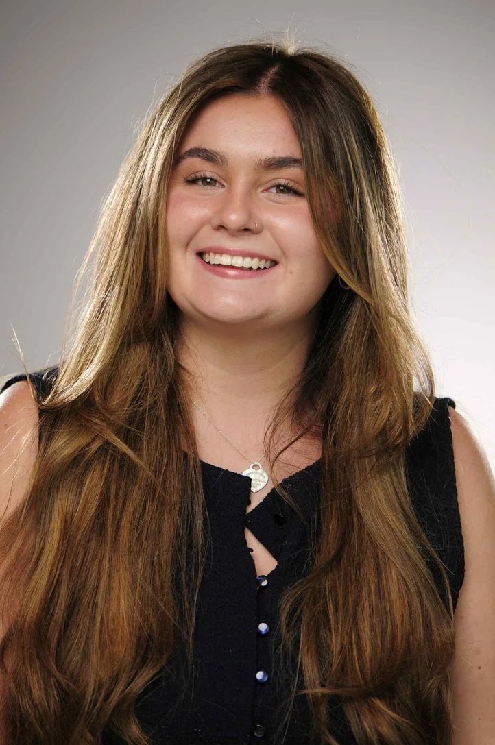 A young woman with long, light brown hair, smiling, wearing a sleeveless black top and a silver heart-shaped pendant necklace.