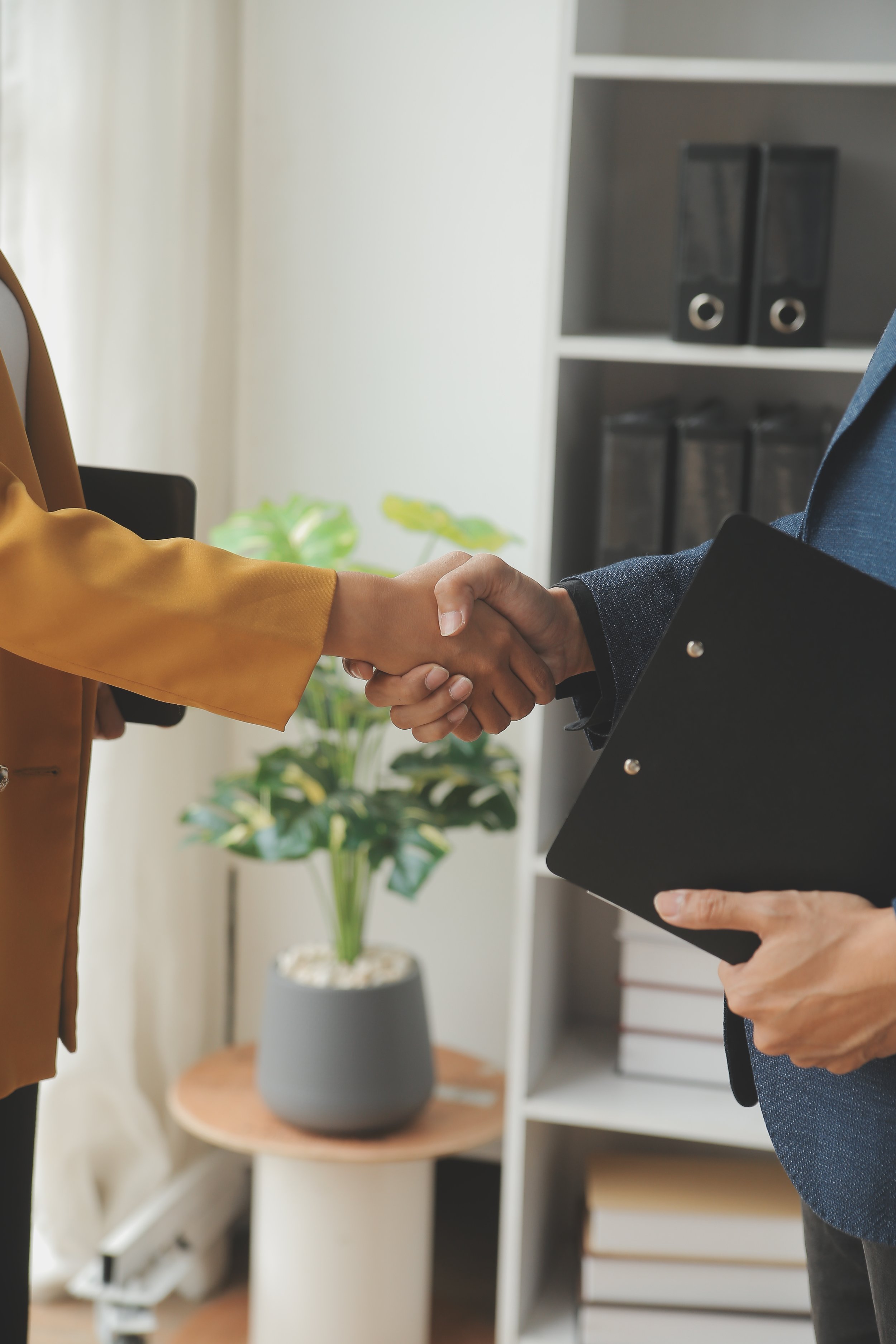 Two people shaking hands in an office setting, one holding a folder, with bookshelves and a potted plant in the background.