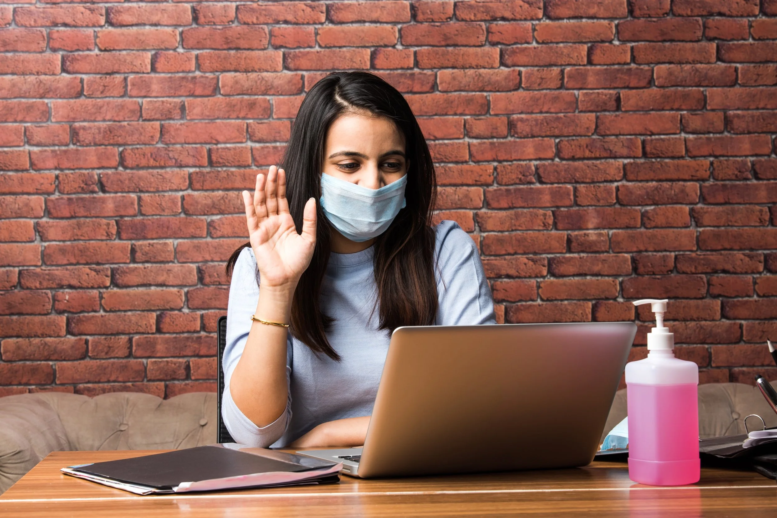 A young woman with long dark hair wearing a light blue face mask, sitting at a desk with a laptop, waving hello or goodbye, with a brick wall in the background, a bottle of hand sanitizer, and some papers.
