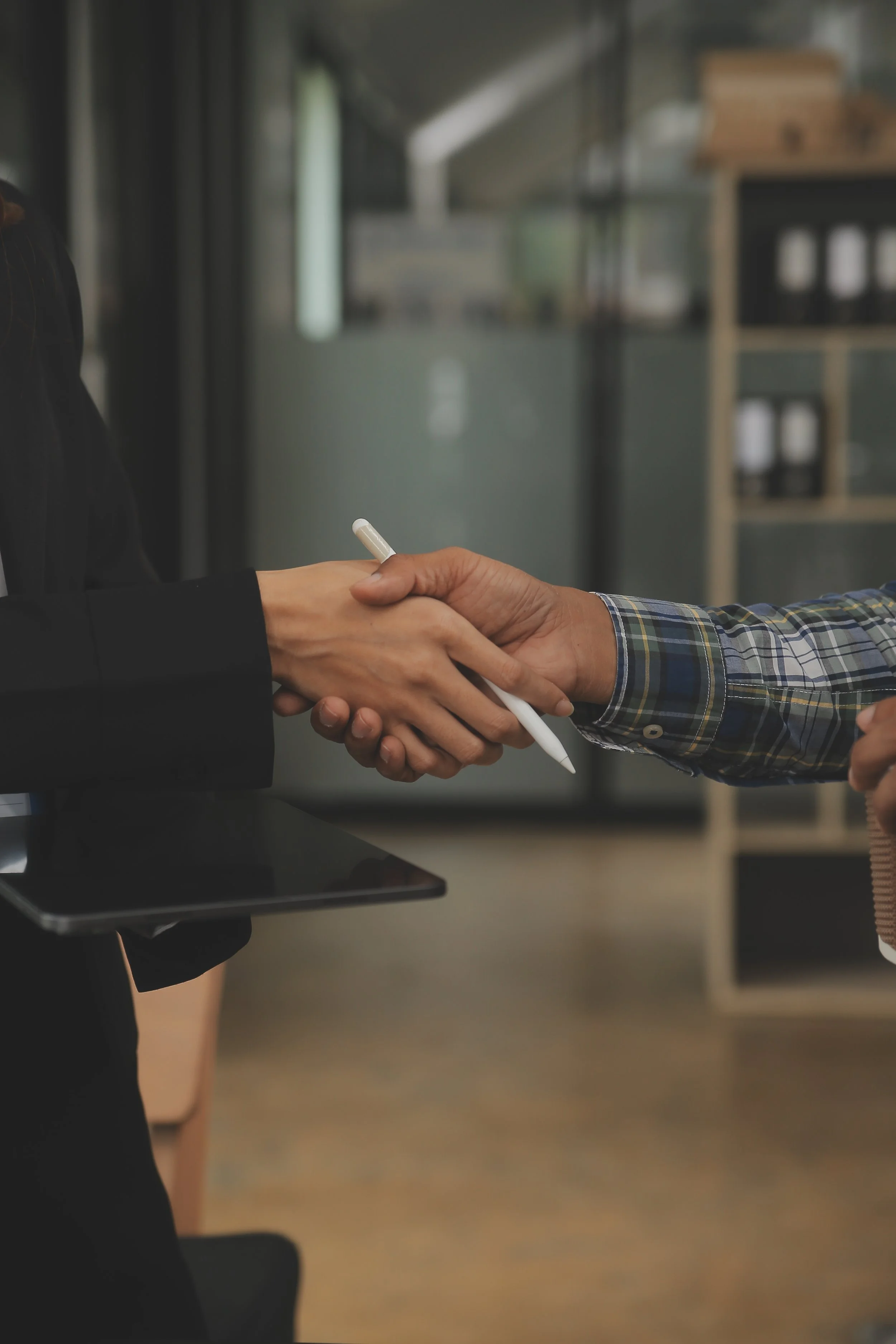 Two people shaking hands, one holding a tablet and a pen, in a professional office setting.