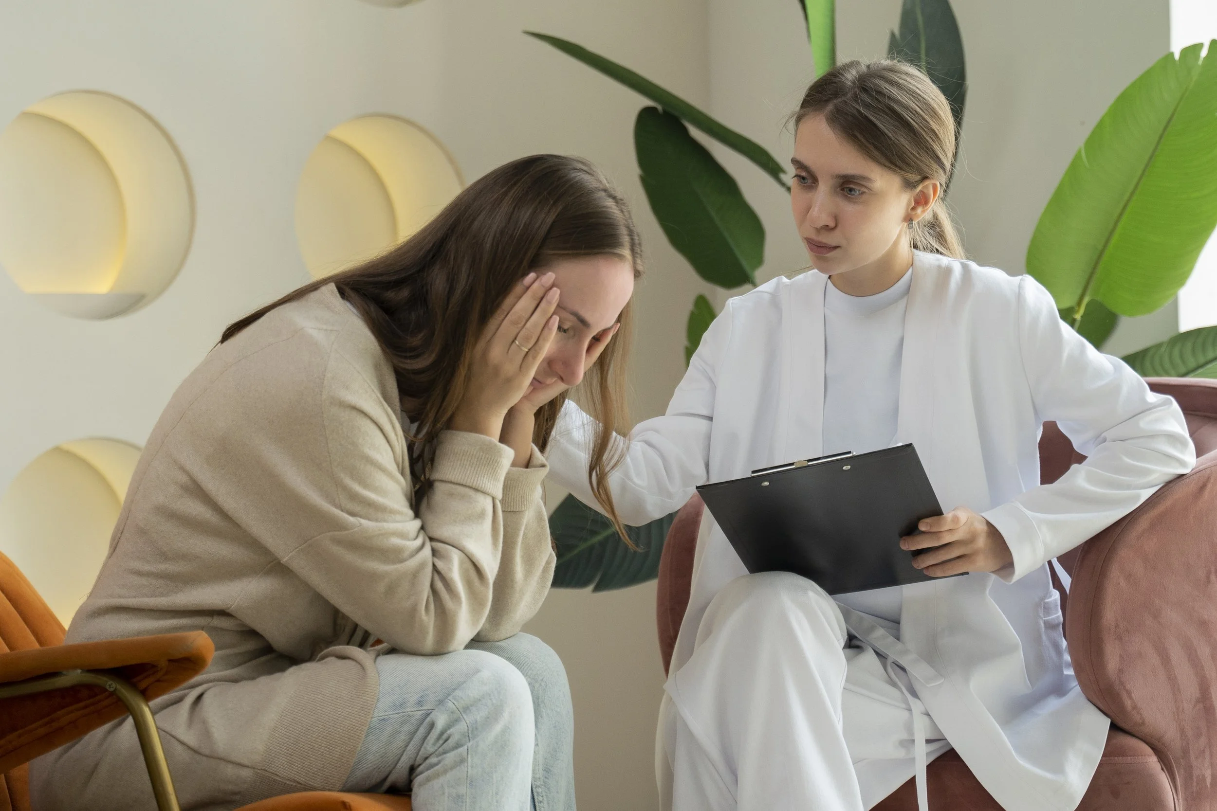 A woman distressed, sitting with her hands on her face, and a female medical professional holding a clipboard, offering comfort and support.