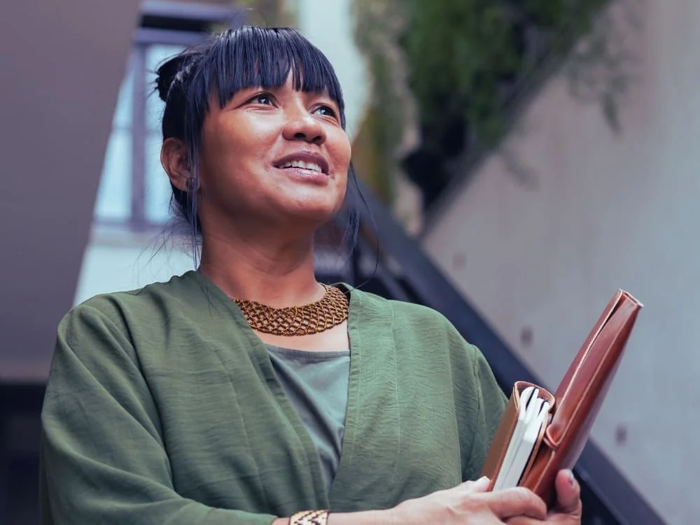 A woman with dark hair, styled with bangs and a bun, is smiling and holding a folder and some notebooks. She is wearing a green jacket, a patterned necklace, and has a positive expression, standing indoors with a staircase and some greenery in the background.