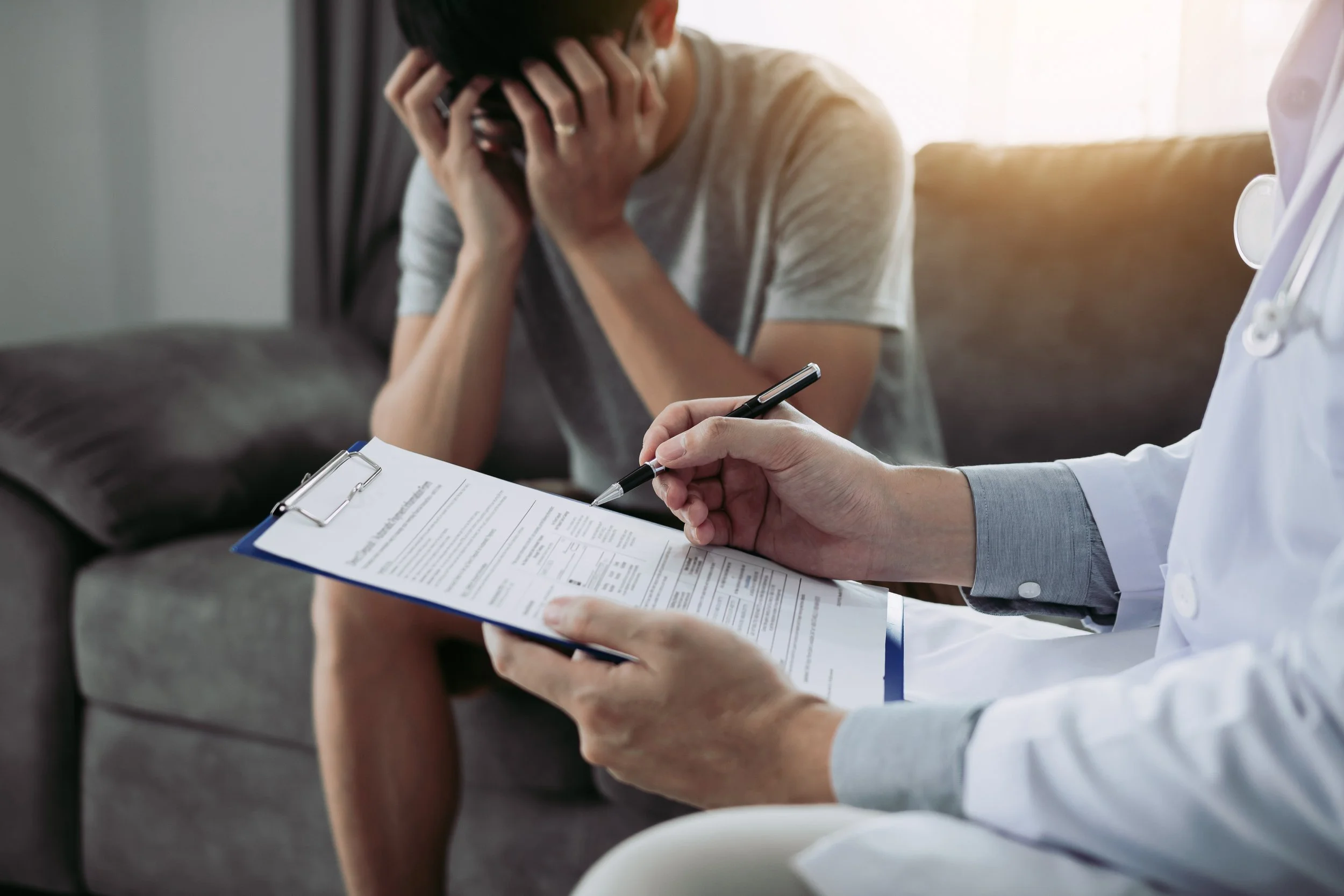 A doctor sitting with a clipboard and pen, talking to a distressed woman with hands on her face during a consultation in a well-lit room.