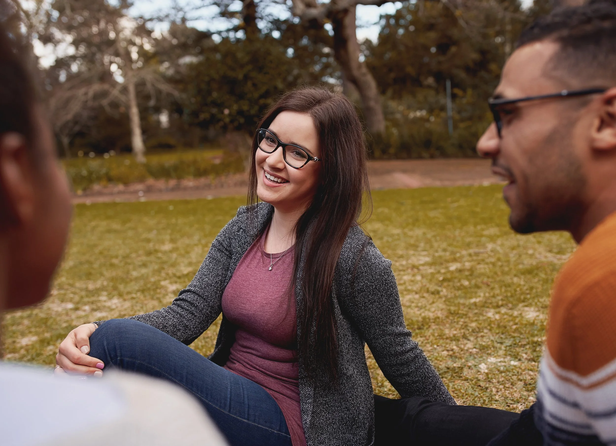 A group of young adults sitting outdoors on grass, talking and smiling, with trees in the background.