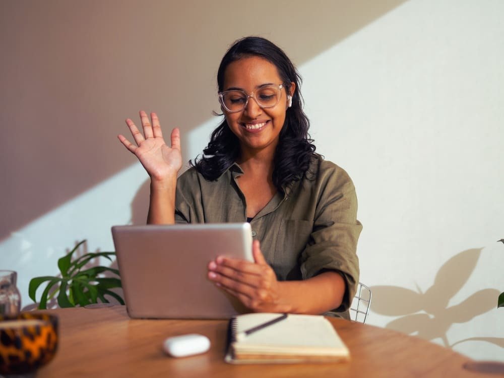 A woman wearing glasses, smiling and waving at a tablet at a desk with a notebook, pen, and some plants.