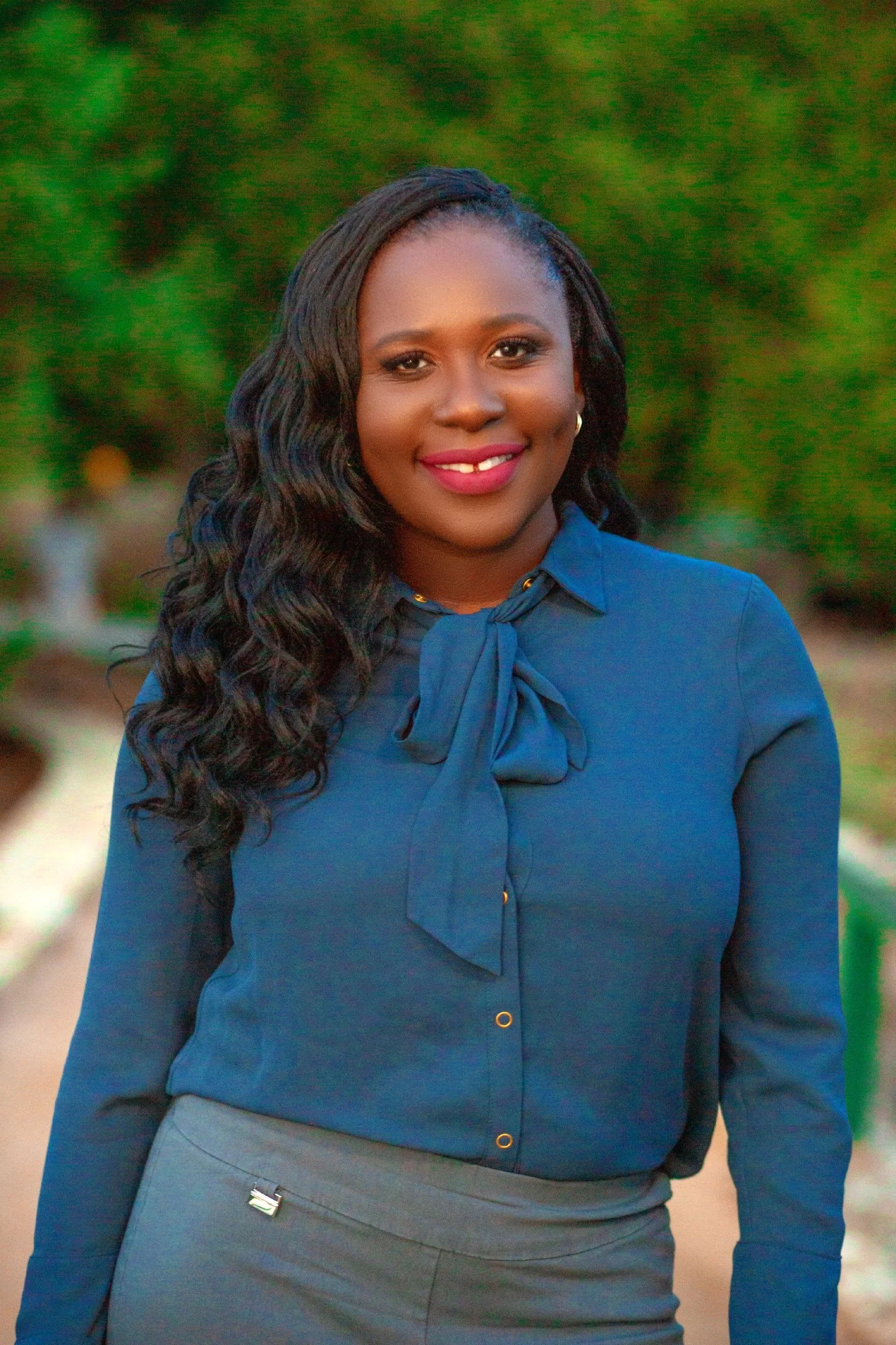 A smiling woman with long, curly black hair, wearing a navy blue blouse with a bow tie at the collar, and gray pants, standing outdoors with green foliage in the background.